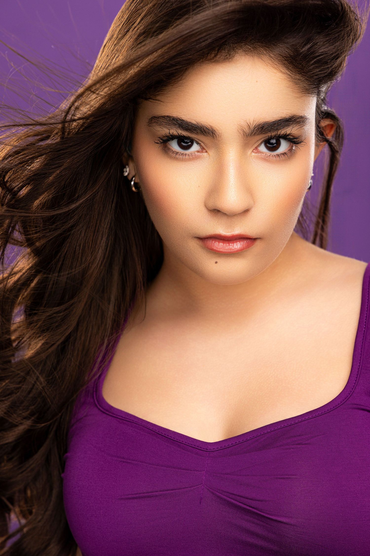 Portrait against purple background wearing dark purple top with dramatic lighting and styled dark hair.