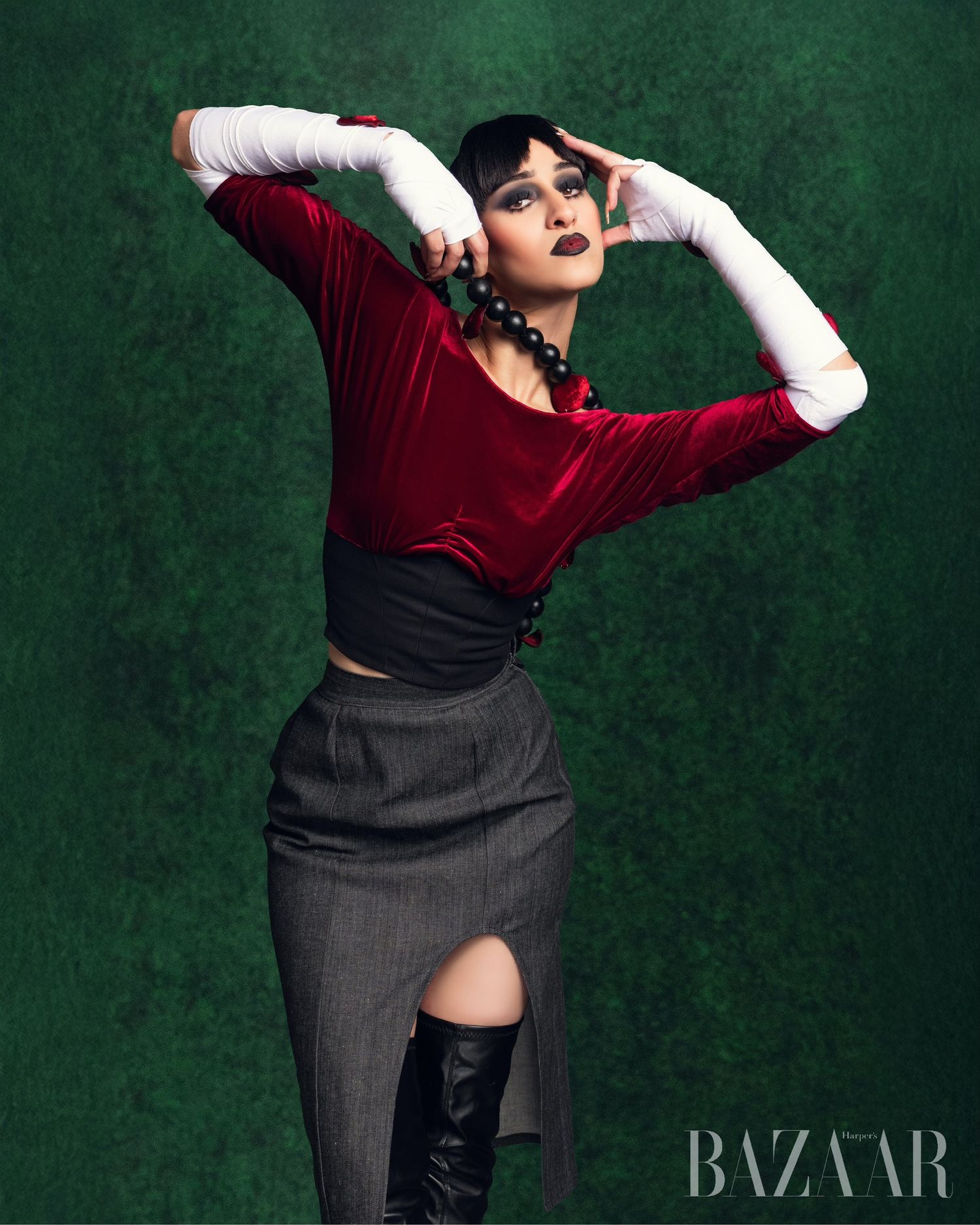 Fashion model in red velvet top and gray skirt with white gloves poses dramatically against dark green background.