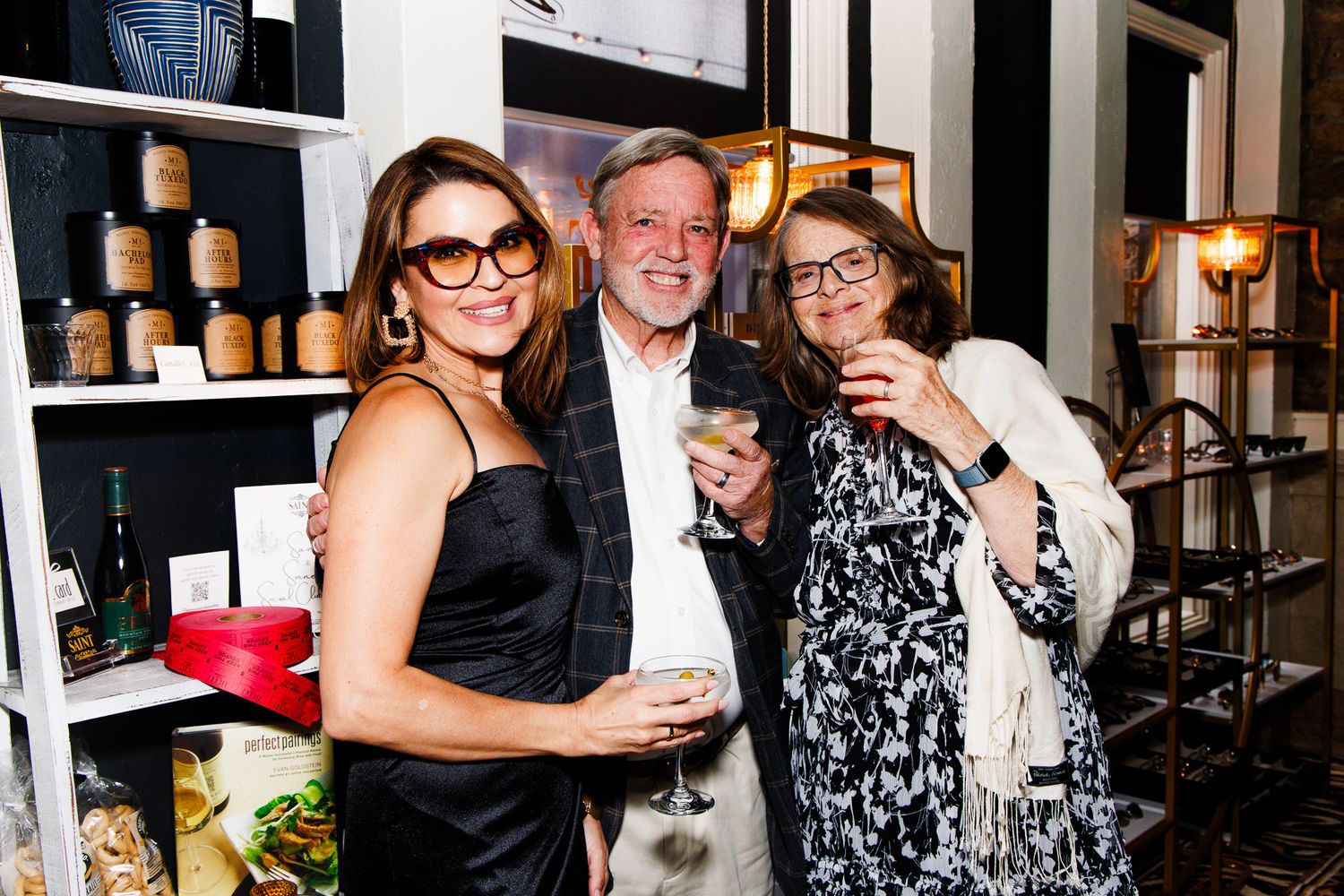 Three friends smile and raise glasses at an elegant indoor social gathering near a well-stocked shelf display.