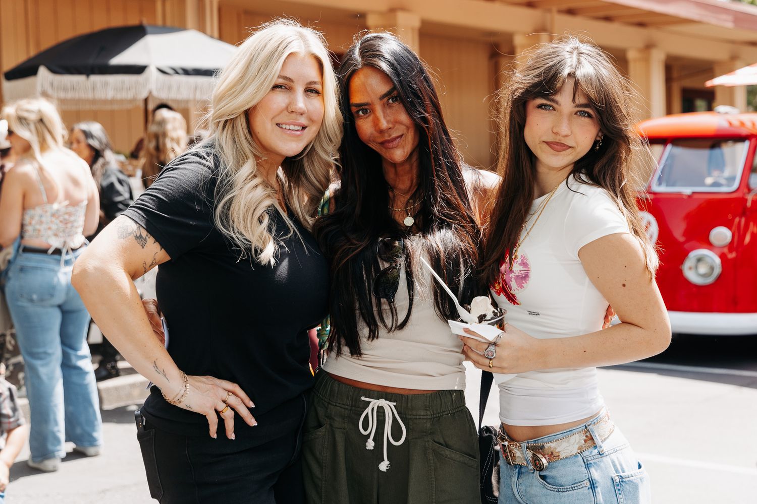 Three women posing together outdoors at a sunny outdoor event near a vintage red vehicle.