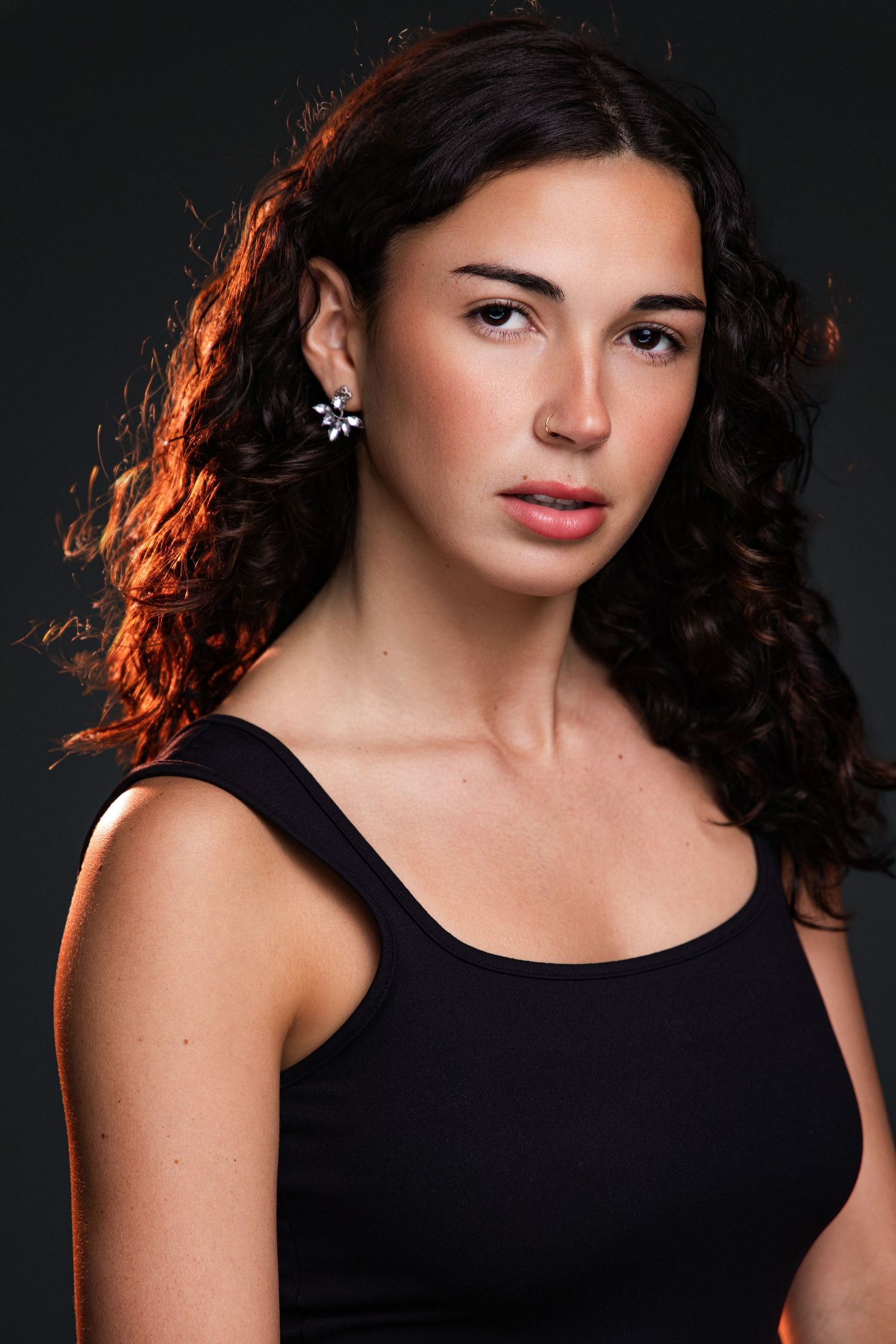 Young woman with curly dark hair wearing a black tank top and floral earring poses against a dark background.