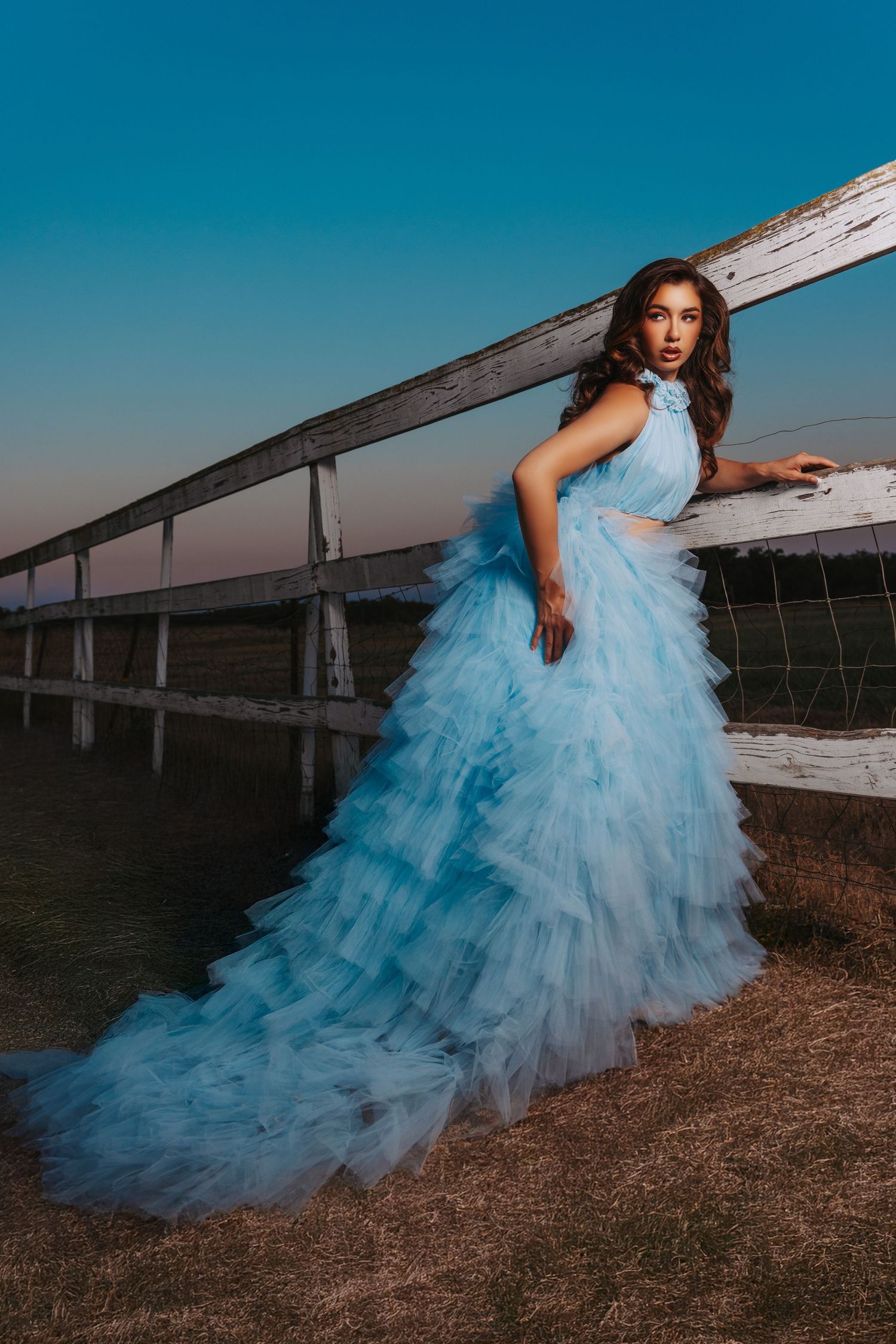 A person in a flowing light blue tulle gown poses dramatically near a wooden boardwalk at sunset.