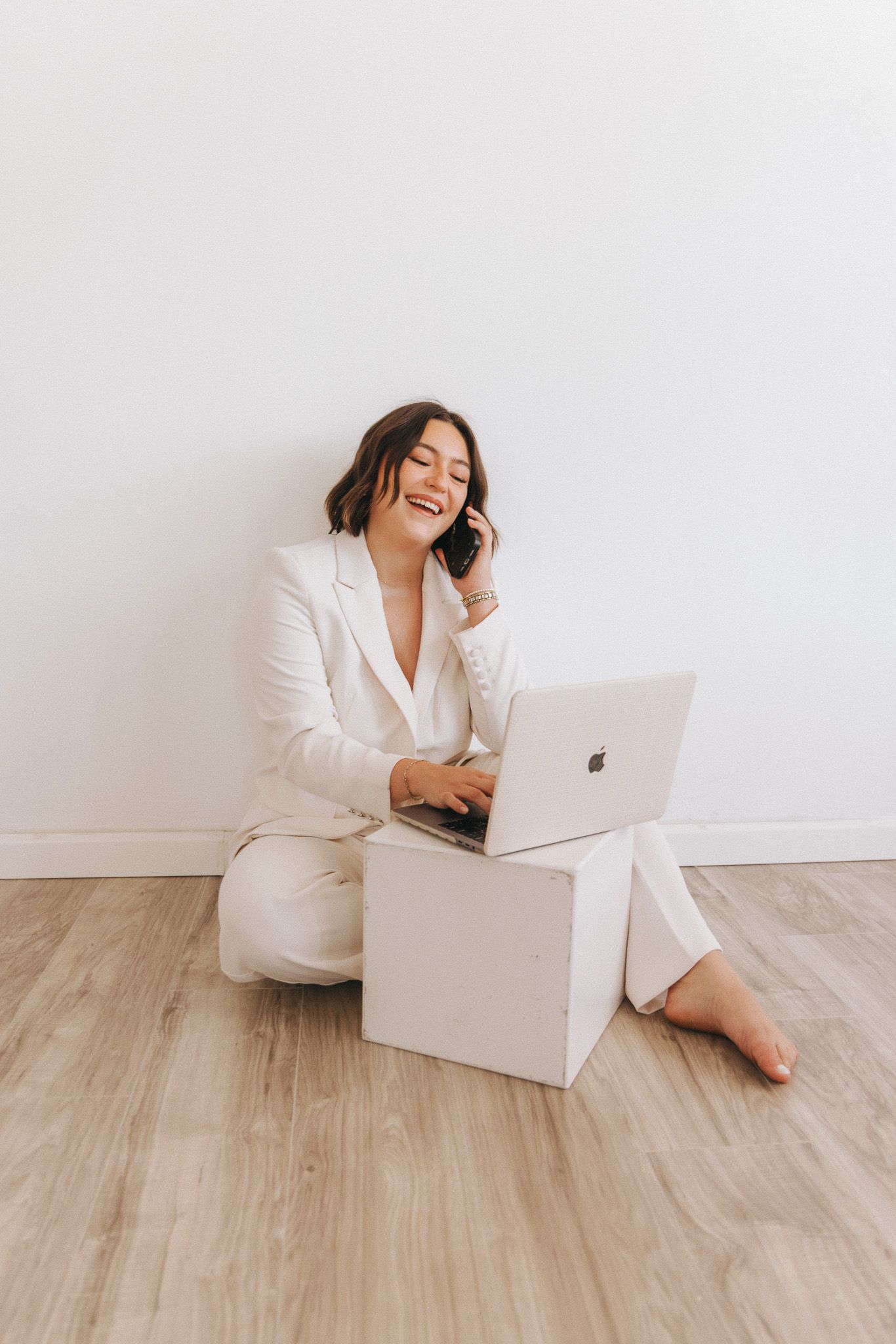 Professional wearing white suit reclines and laughs while working remotely on a laptop from a minimalist white room.