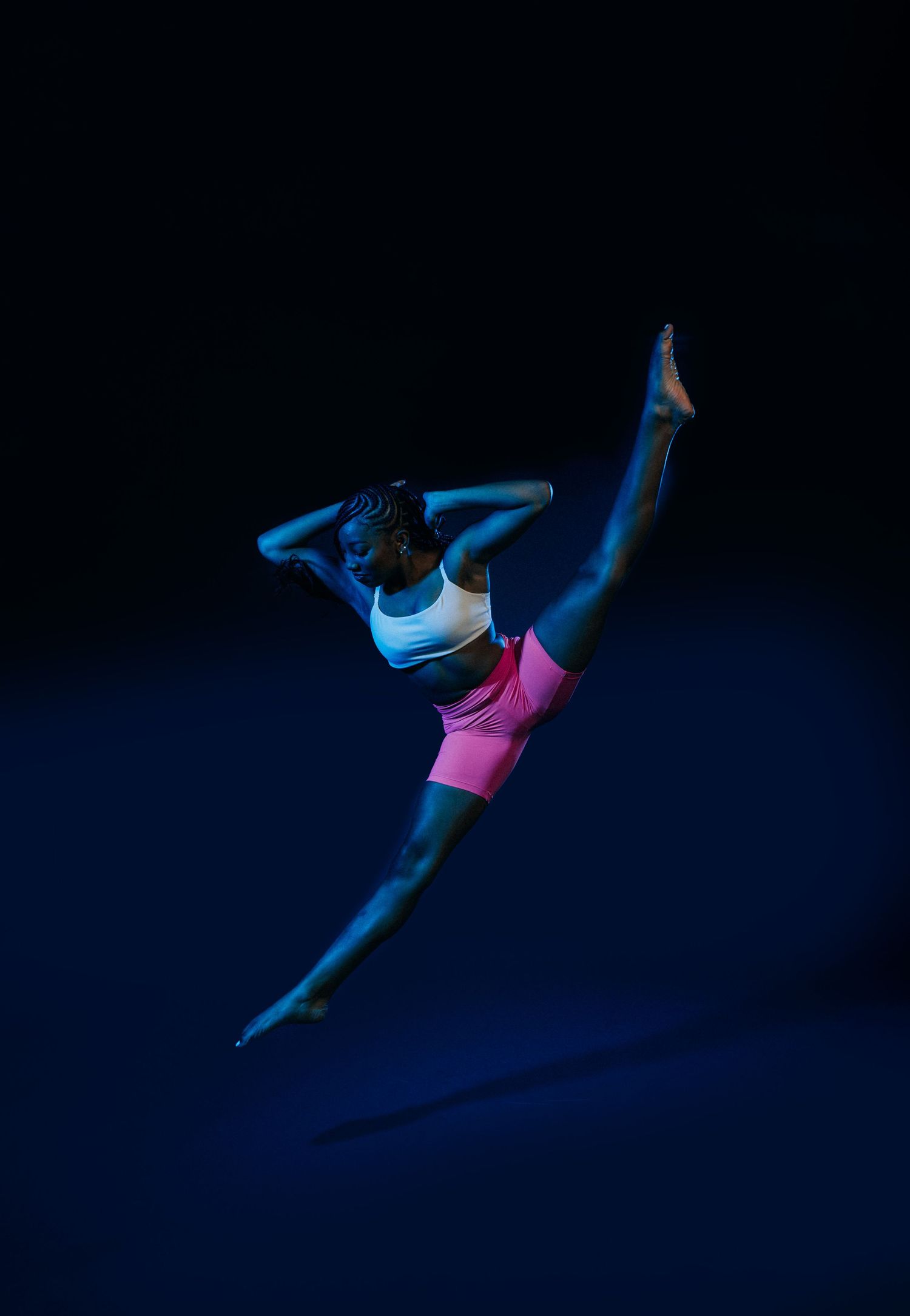 Graceful dancer leaps through air in athletic pose against dark background with blue lighting effect.