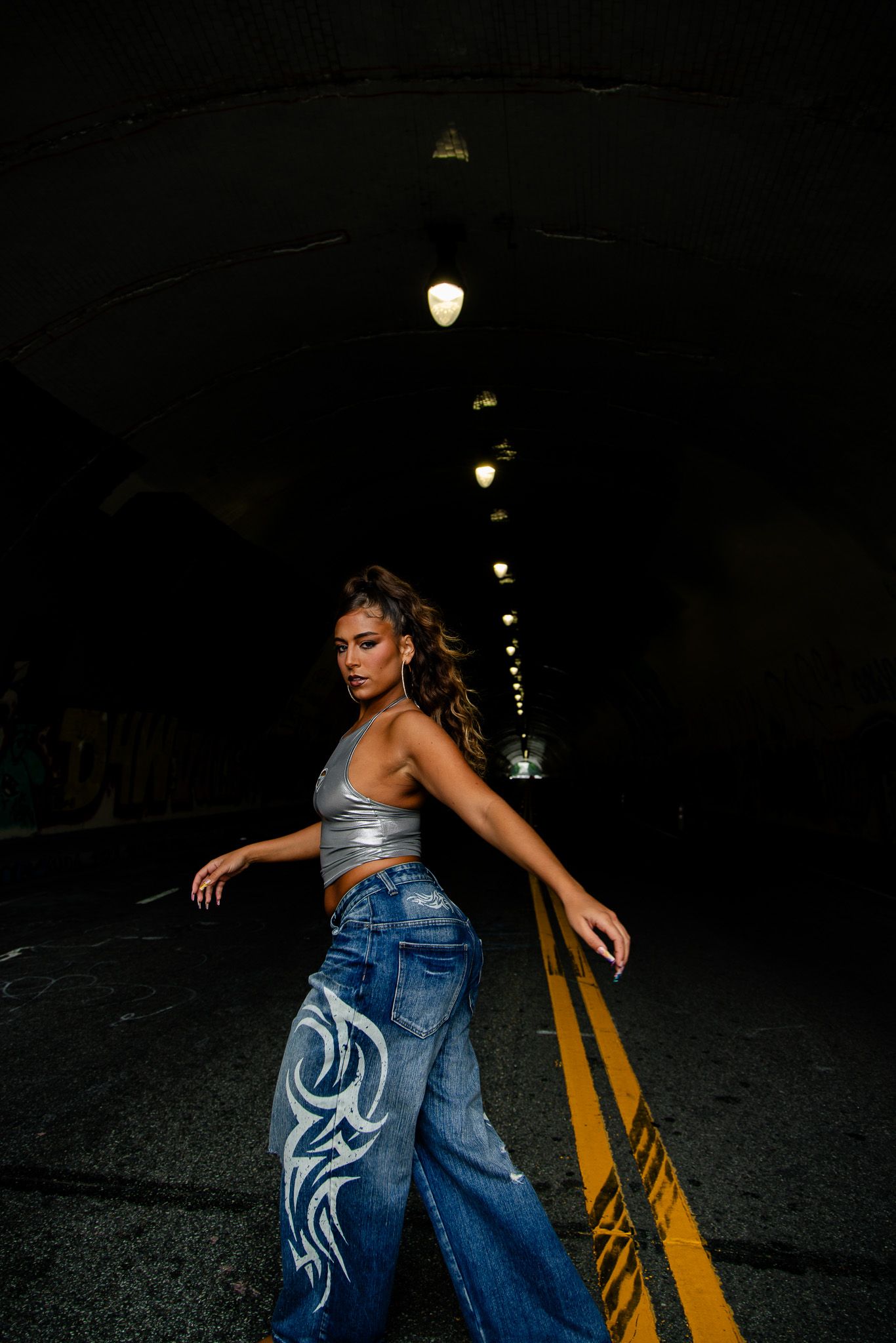Person in crop top and painted jeans dances on dark street with yellow lines and streetlights in background.