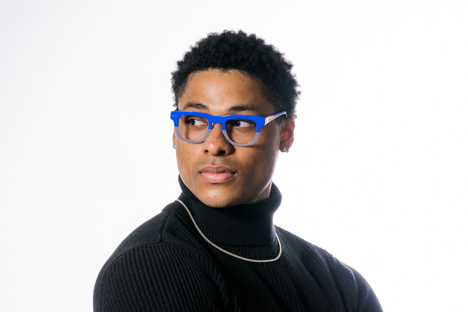 Young man wearing bold blue rectangular glasses and black turtleneck with gold chain against white background.