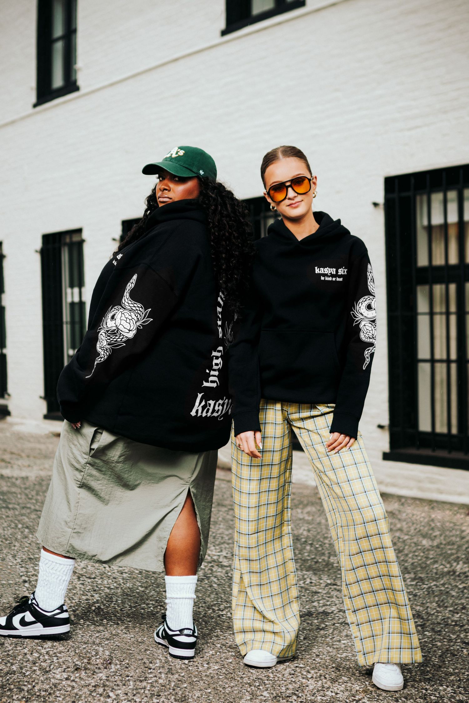 Two women posing outdoors in matching black graphic hoodies, styled with plaid pants, skirt, sneakers, and accessories.