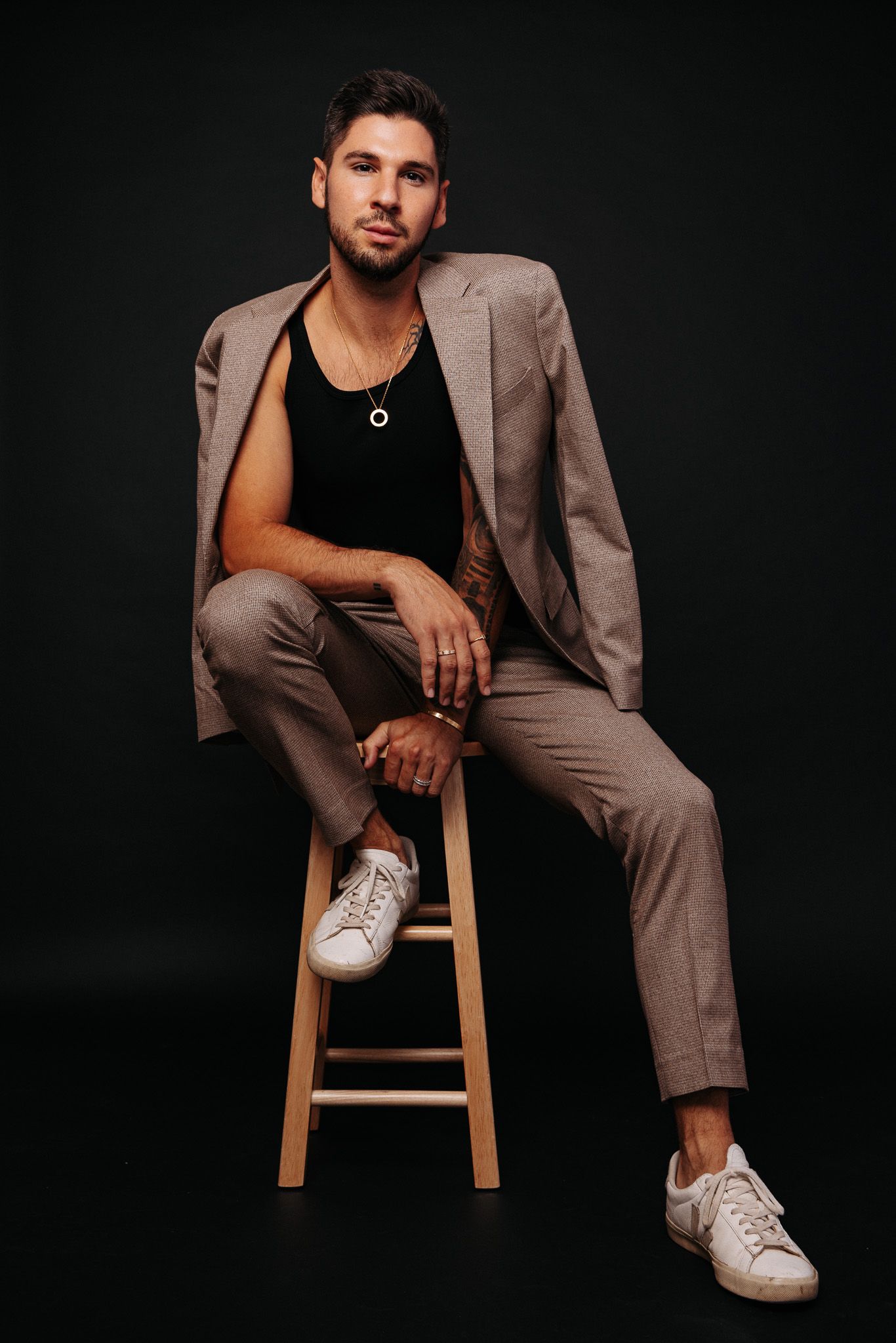 Fashion portrait against dark background showing model in beige suit and black tank top sitting on wooden stool.