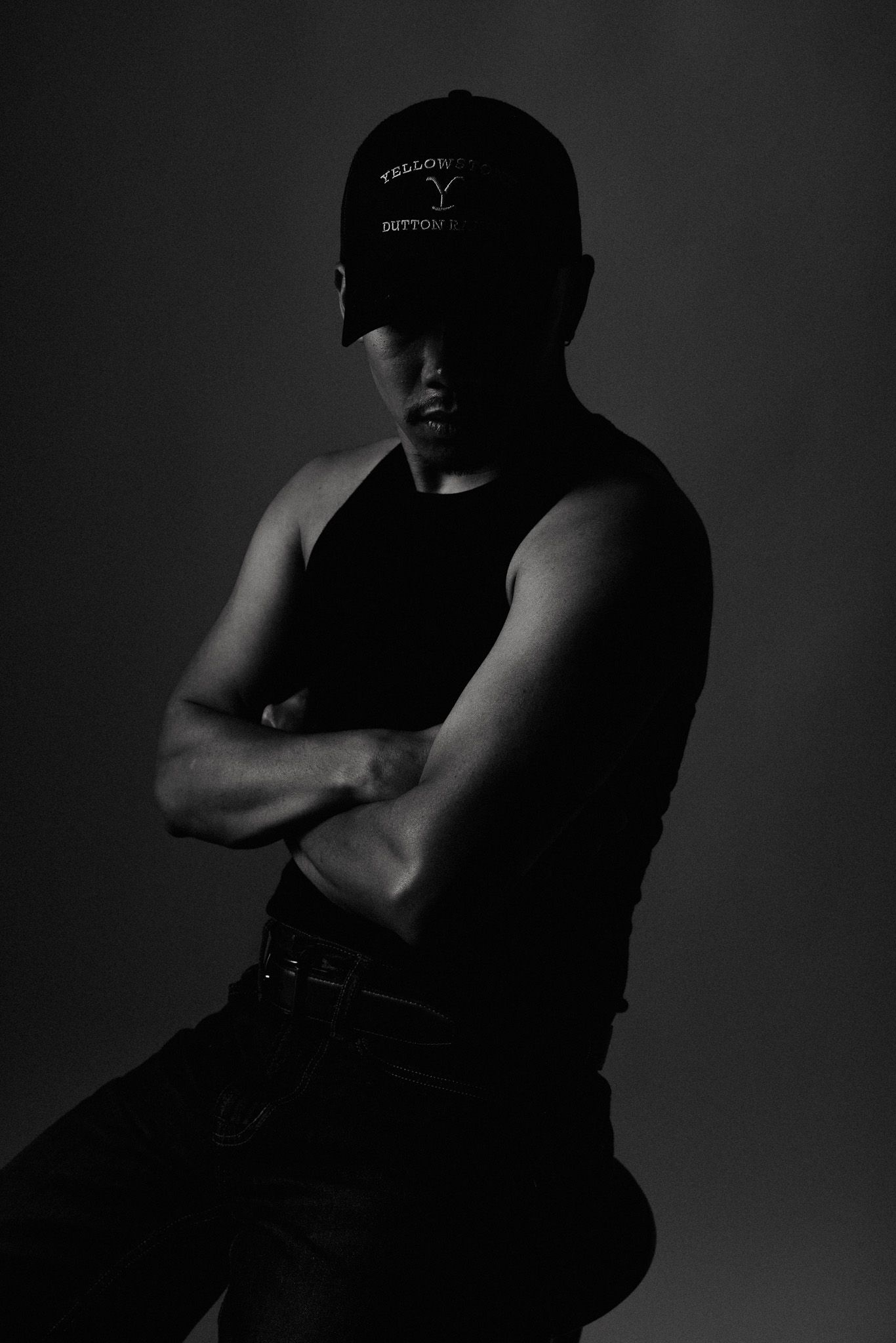 Dramatic black and white portrait of someone in a baseball cap and tank top with crossed arms against dark backdrop.