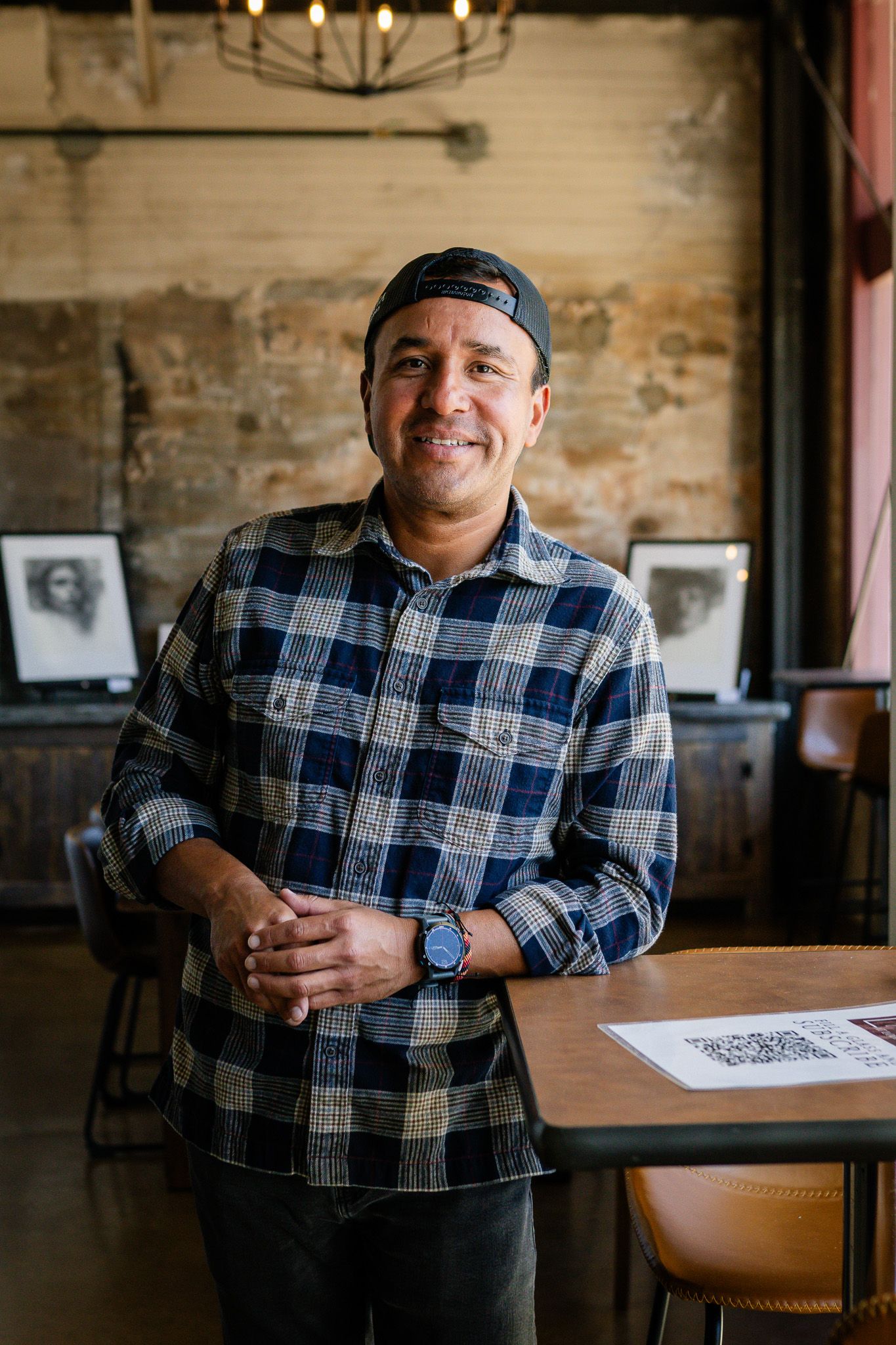 Restaurant worker in plaid flannel shirt and backwards cap stands smiling in casual dining establishment.