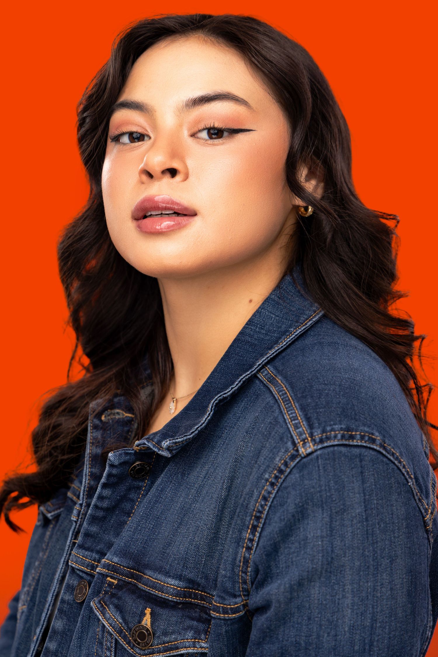Young model wearing denim jacket poses confidently against vibrant orange background.