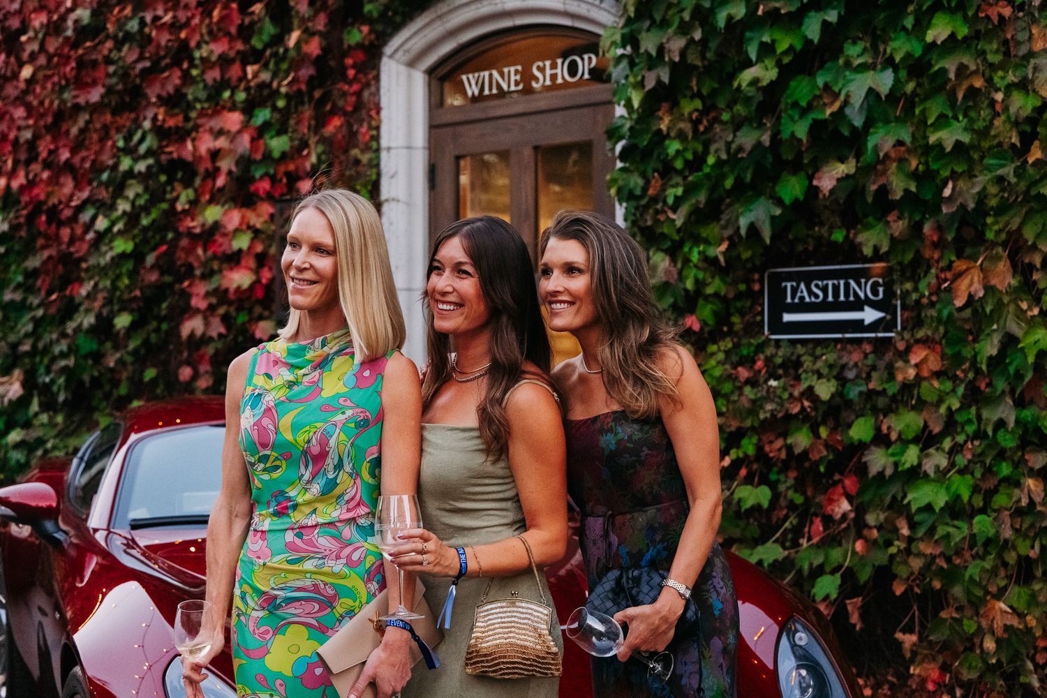 Three women in summer dresses walk together past an ivy-covered building wall on a sunny day.