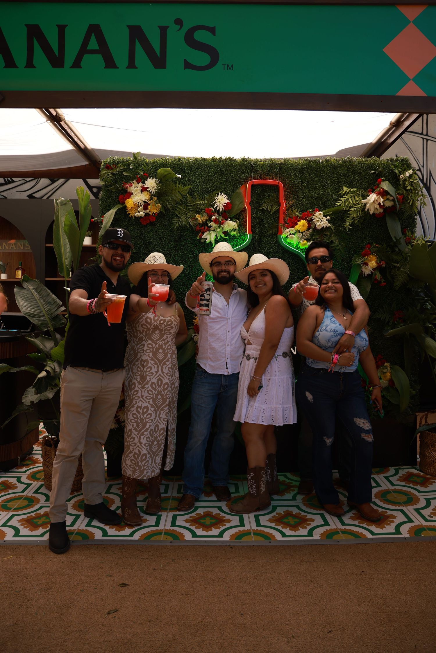 Group of friends in cowboy hats toasting drinks at Buchanan's branded outdoor event booth with green plant wall backdrop.