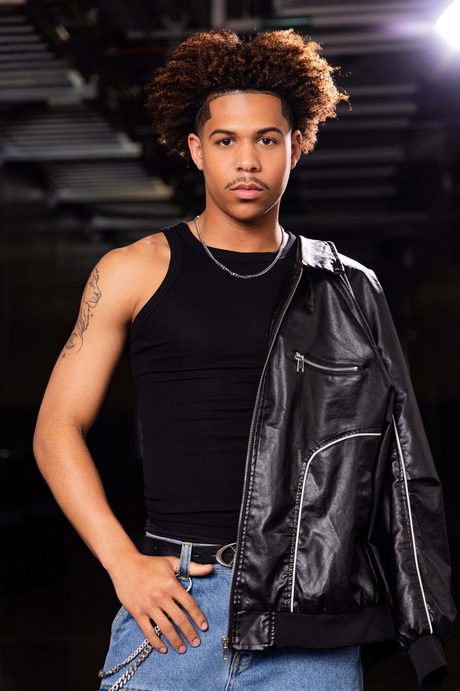 Young man with curly hair poses in black tank top, leather jacket over shoulder, and jeans in a moody studio setting.