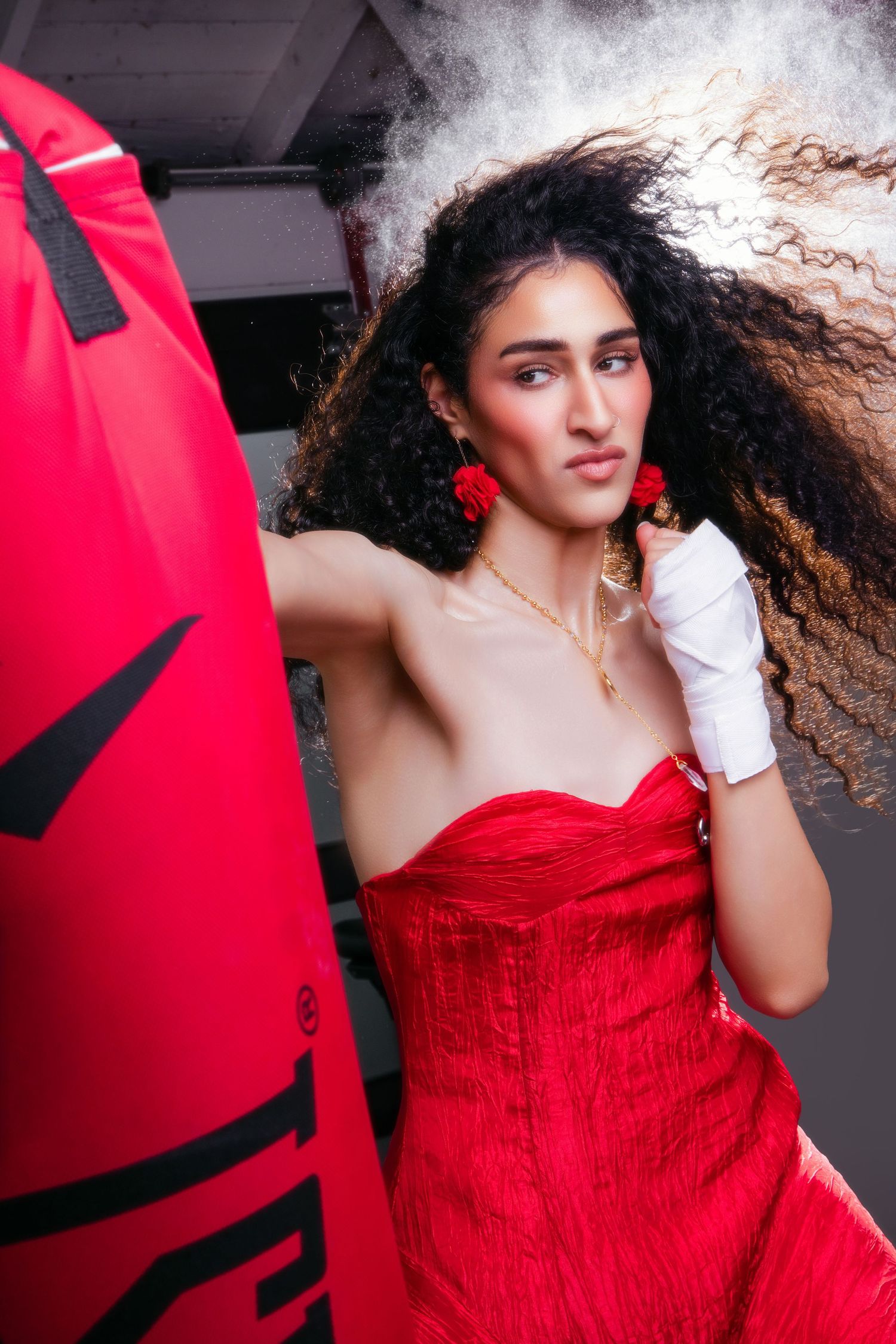 Woman in red strapless dress with white boxing wraps punches a red heavy bag, curly hair flying.