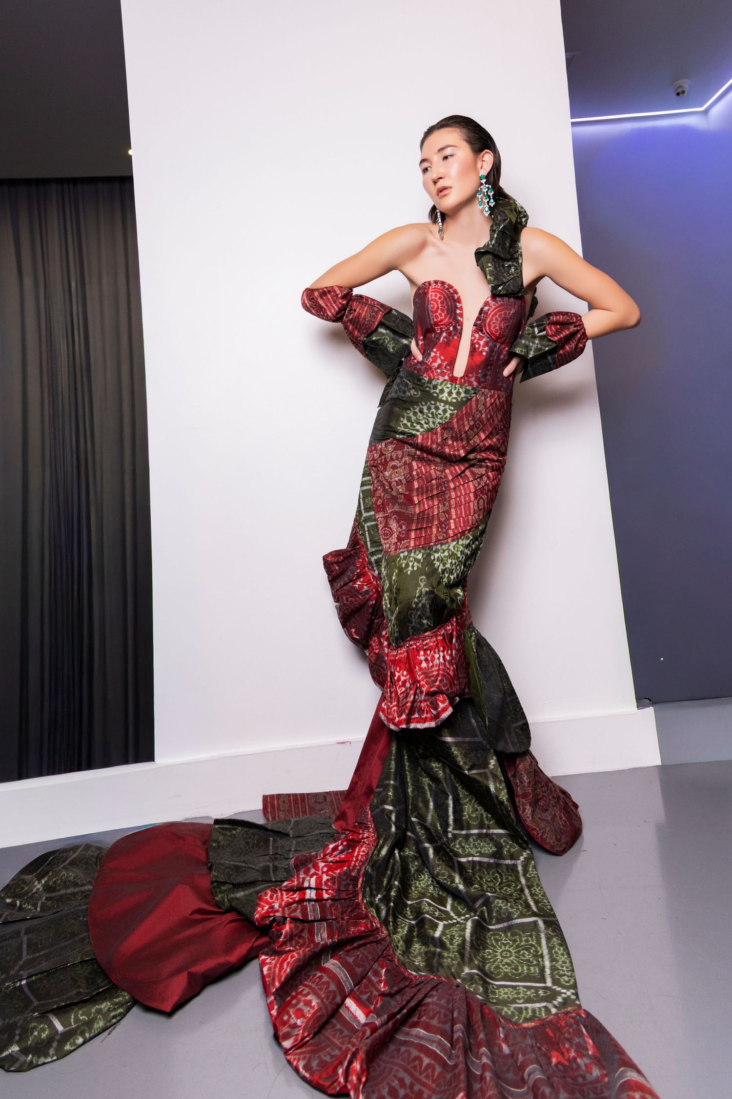 Fashion model poses in dramatic red and green patterned evening gown against studio backdrop.