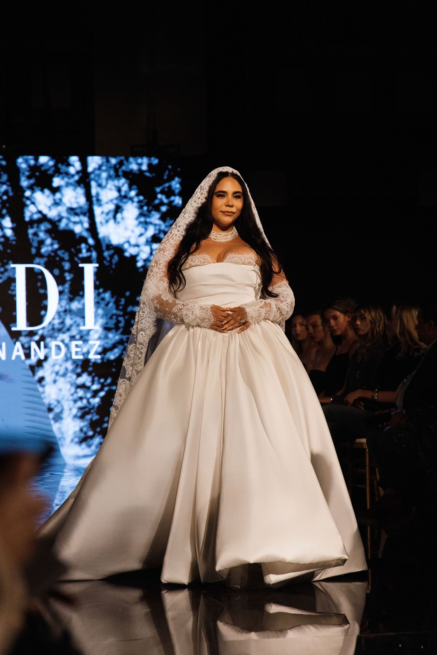Models showcase elegant white wedding gowns with dramatic silhouettes on a dark runway at a bridal fashion show.