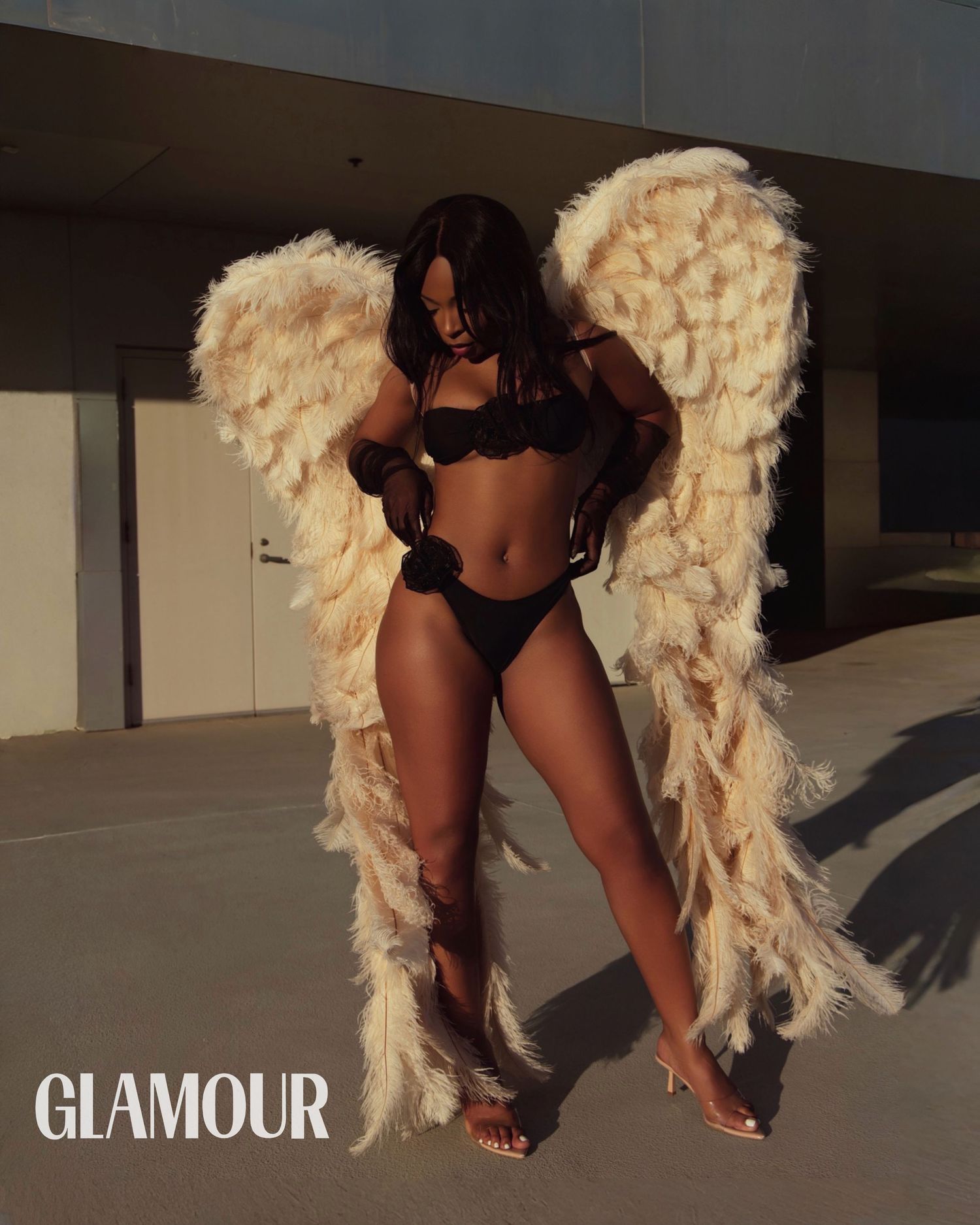 Woman in black bikini poses with large feathered angel wings outdoors in a Glamour magazine photoshoot.