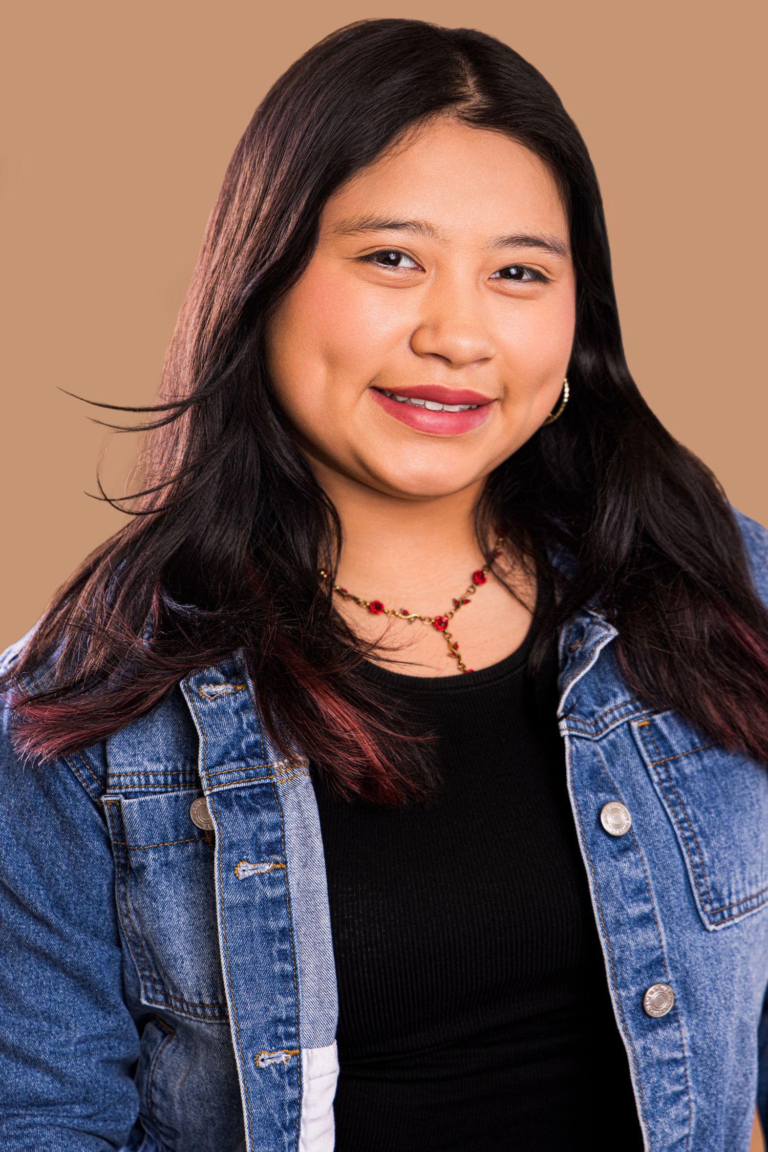 Smiling young woman with long dark hair wearing a denim jacket and red necklace against a tan background.