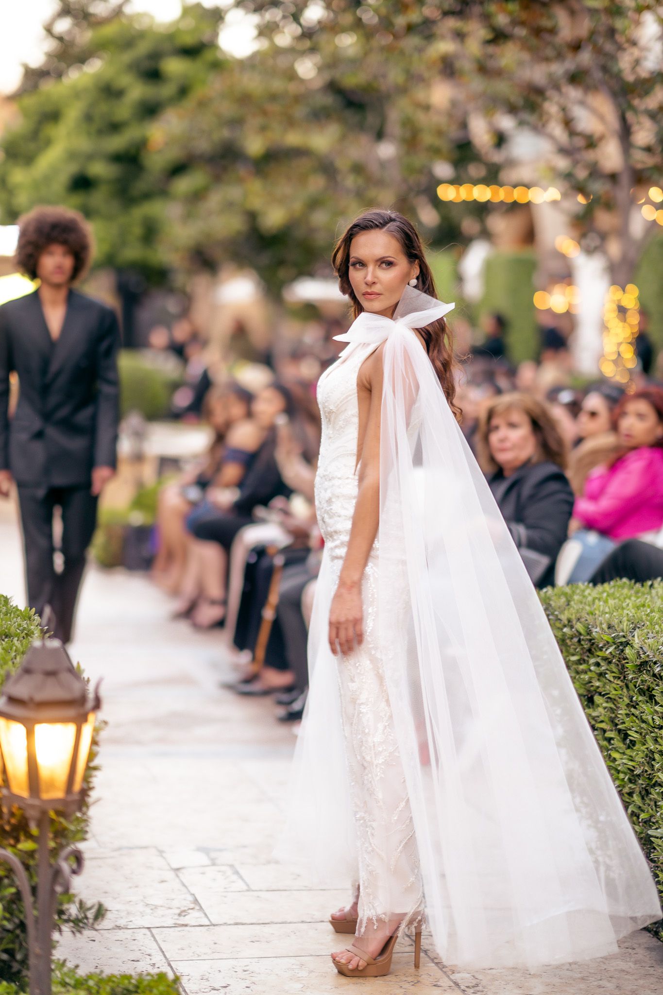 Model wearing an elegant white bridal gown with flowing veil poses on garden path during outdoor fashion show.