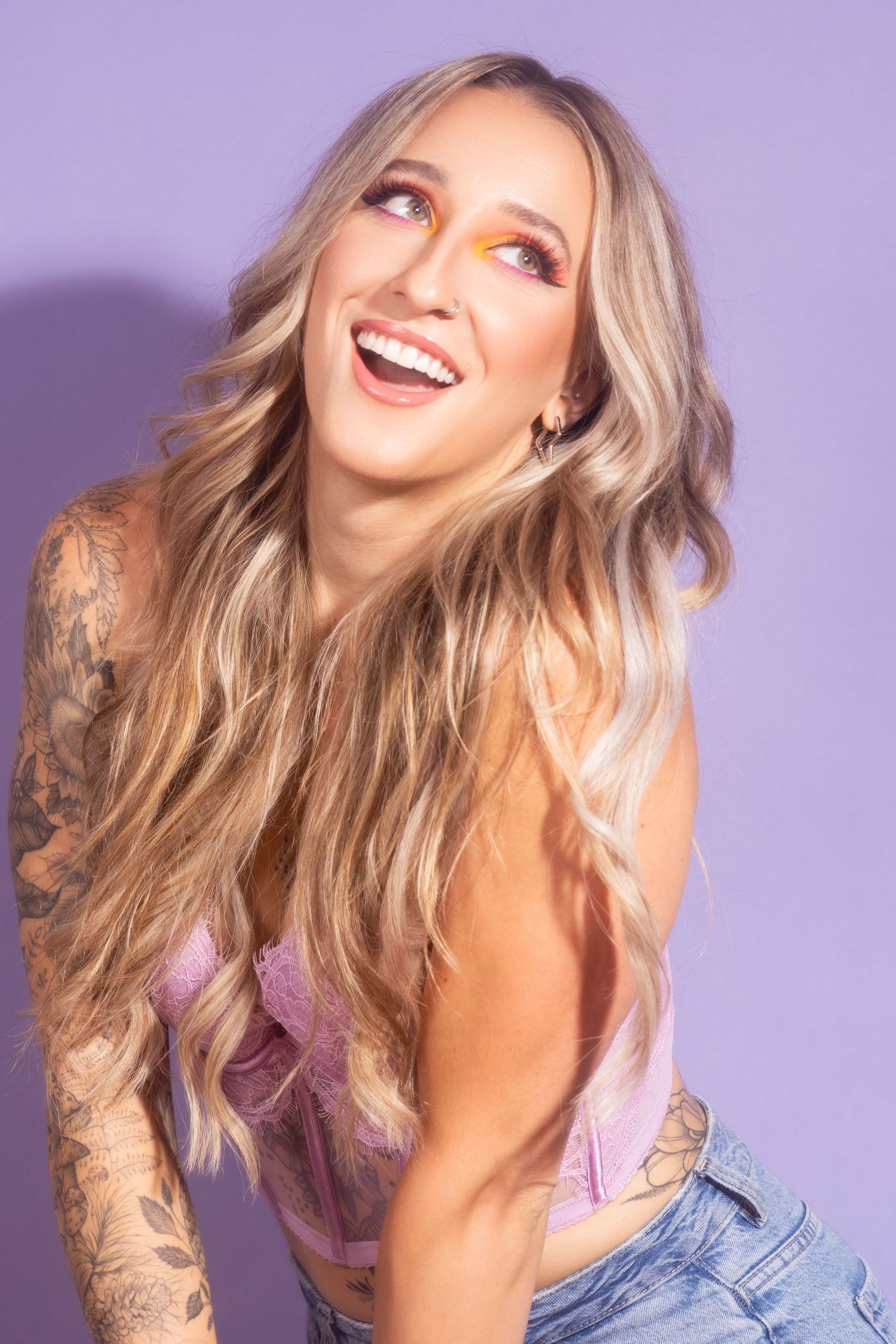 Fashion model with blonde waves wearing denim and pink outfit poses playfully against purple backdrop.
