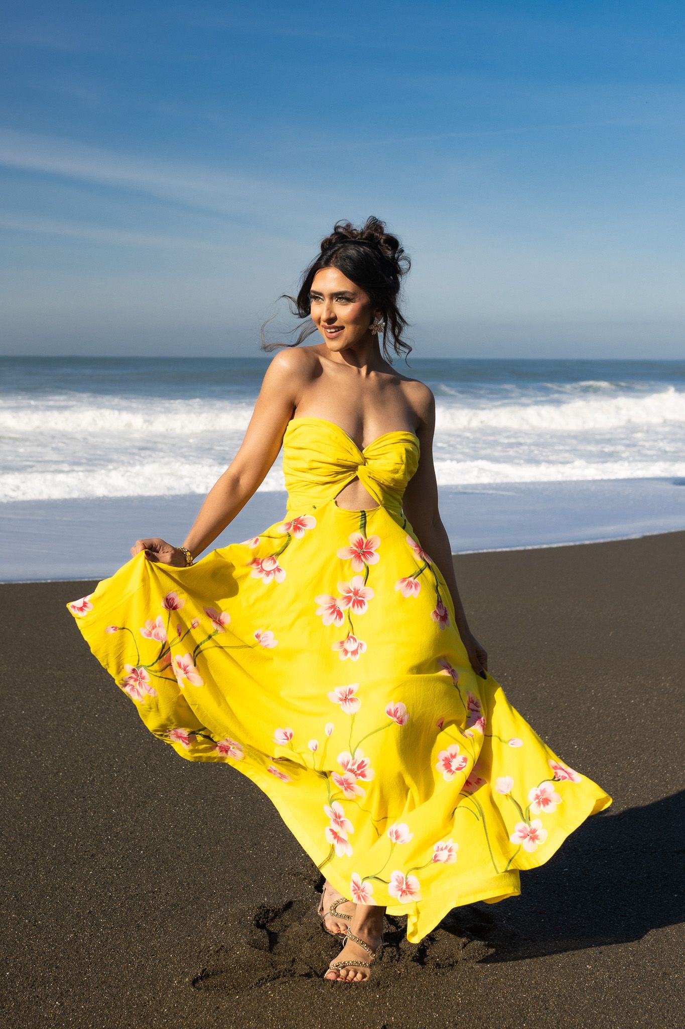 A vibrant yellow floral dress flows in the wind as someone poses playfully on a dark sandy beach by ocean waves.