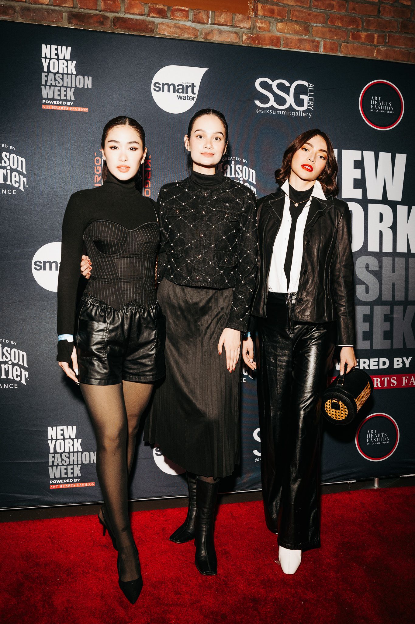 Three fashionable attendees in dark elegant outfits pose at a New York Fashion Week event against a branded backdrop.