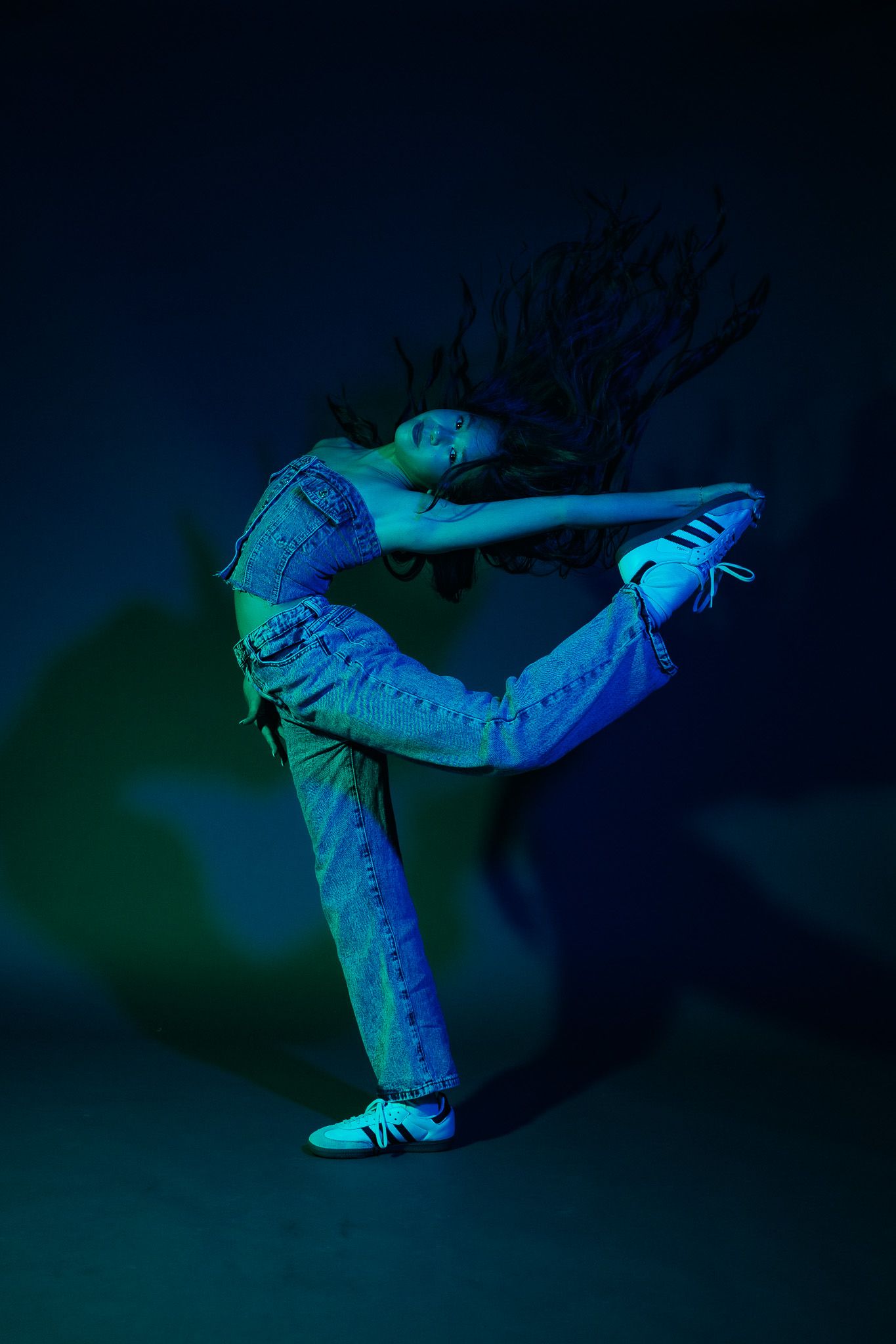 A dancer performs a dynamic pose in blue lighting wearing denim and sneakers, creating an artistic silhouette.