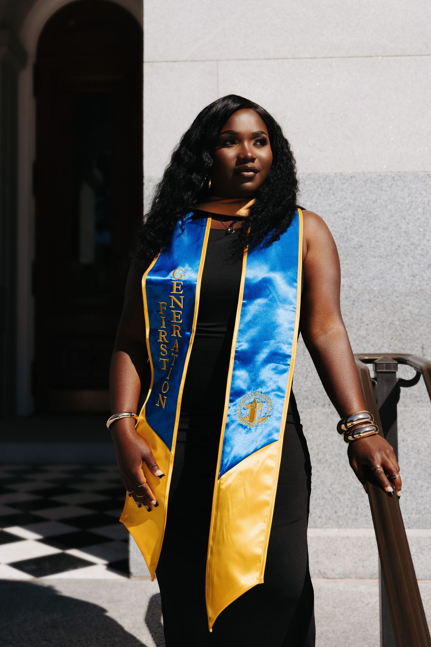 Graduate wearing blue and yellow sash celebrates outdoors in sleeveless black dress next to white wall.