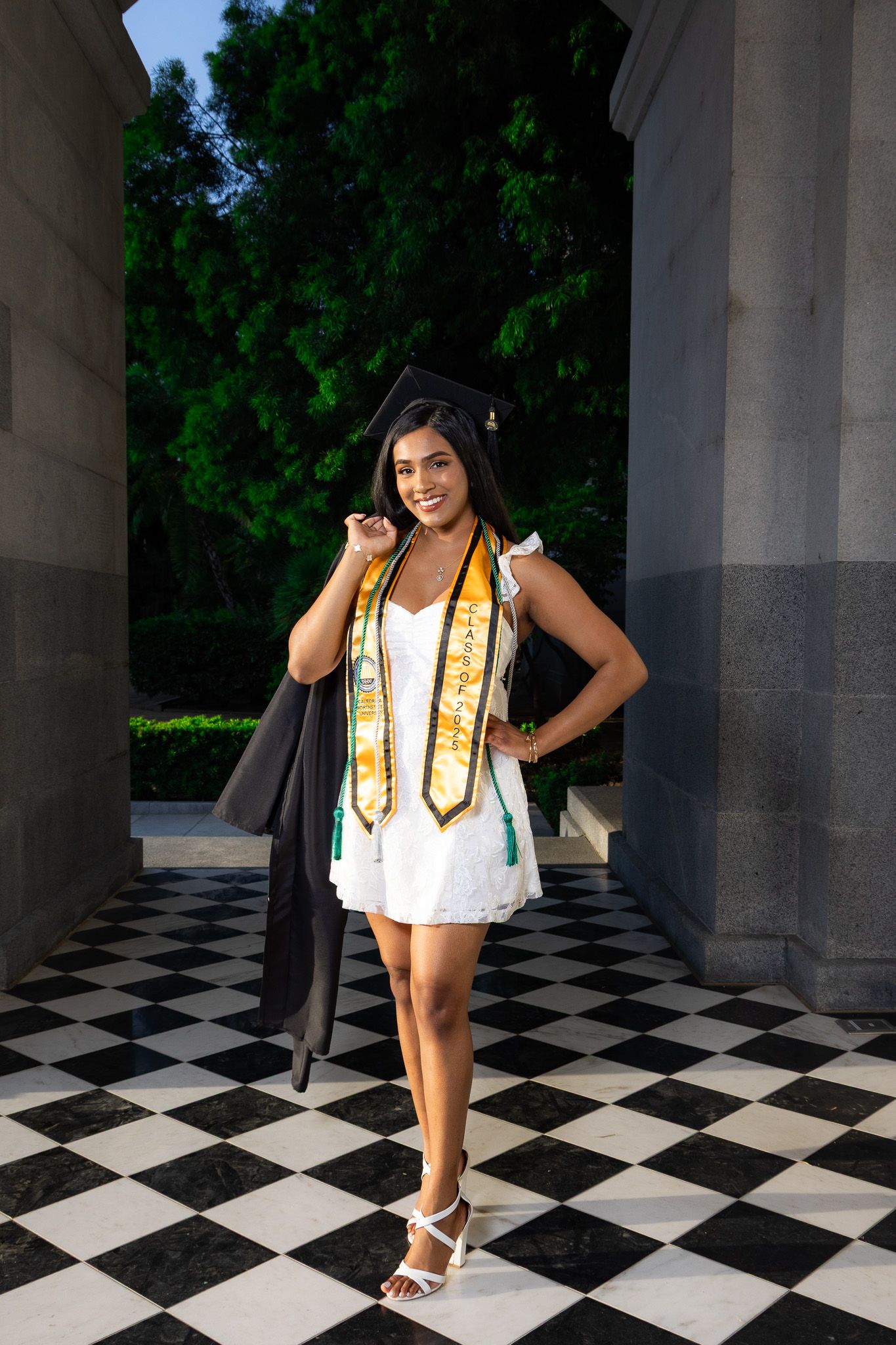 Graduate in white dress with gold leis poses for photos on checkered floor near stone columns outdoors.