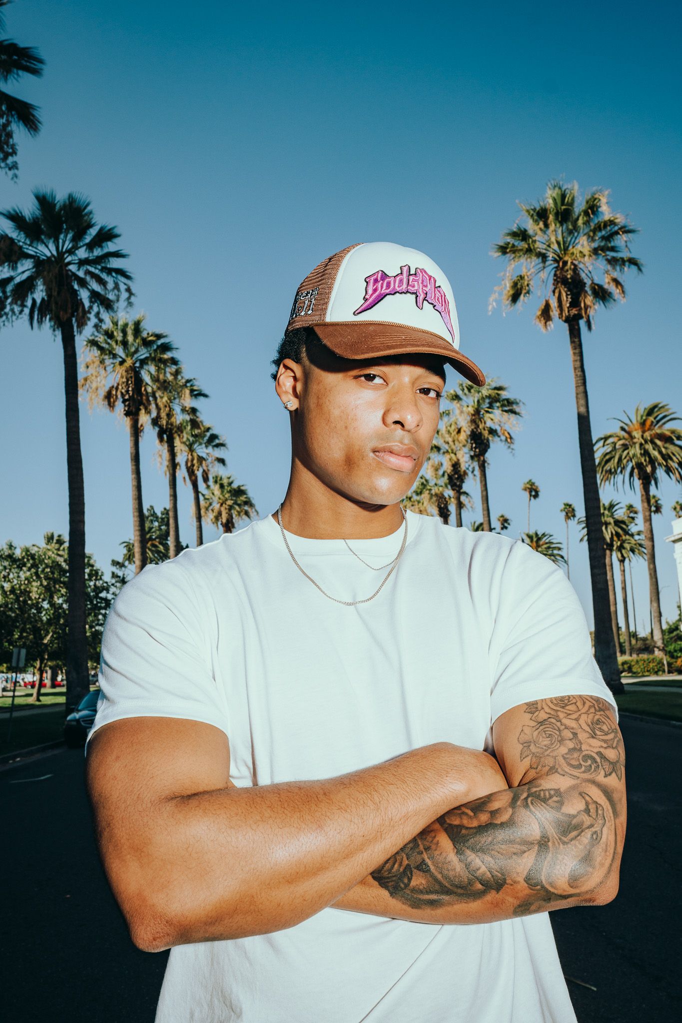 Young man with tattoos and trucker hat poses arms crossed before palm trees in sunny Los Angeles.