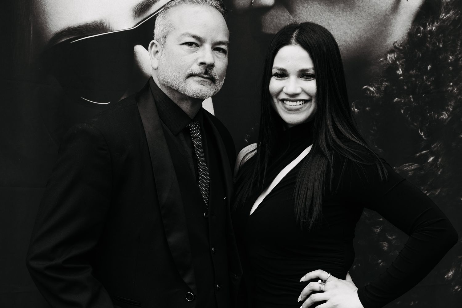 Black and white photo of a smiling man in a suit and woman in black dress posing together at a formal event.