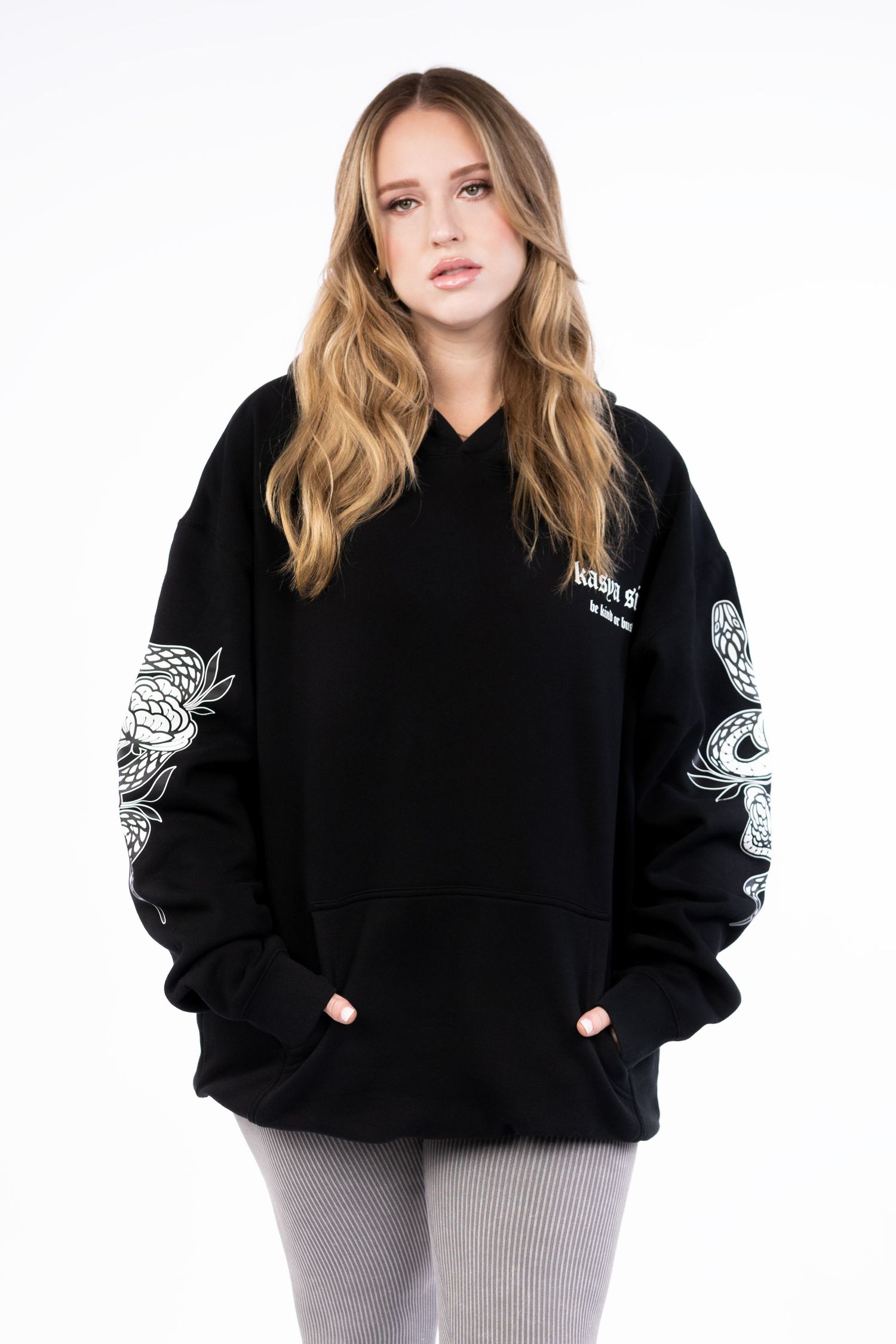 Woman wearing oversized black hoodie with white floral snake sleeve print and kangaroo pocket, paired with grey leggings.