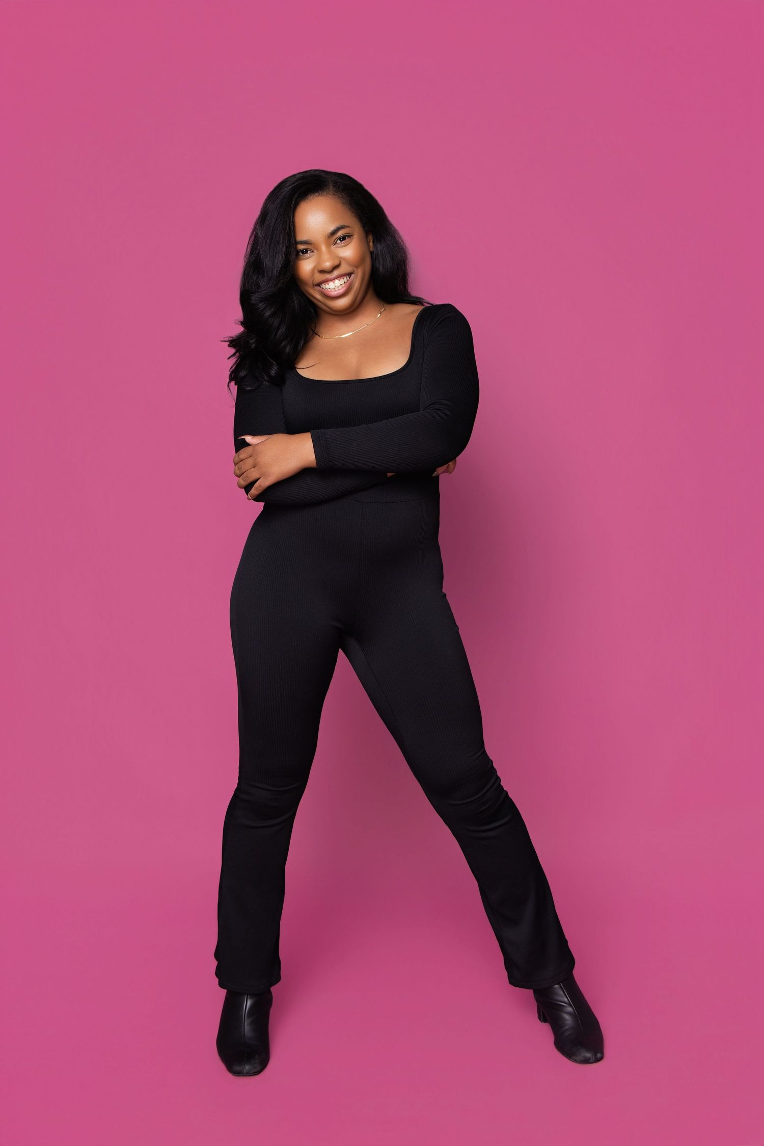 Smiling woman in black jumpsuit and boots posing confidently against a vibrant pink background.