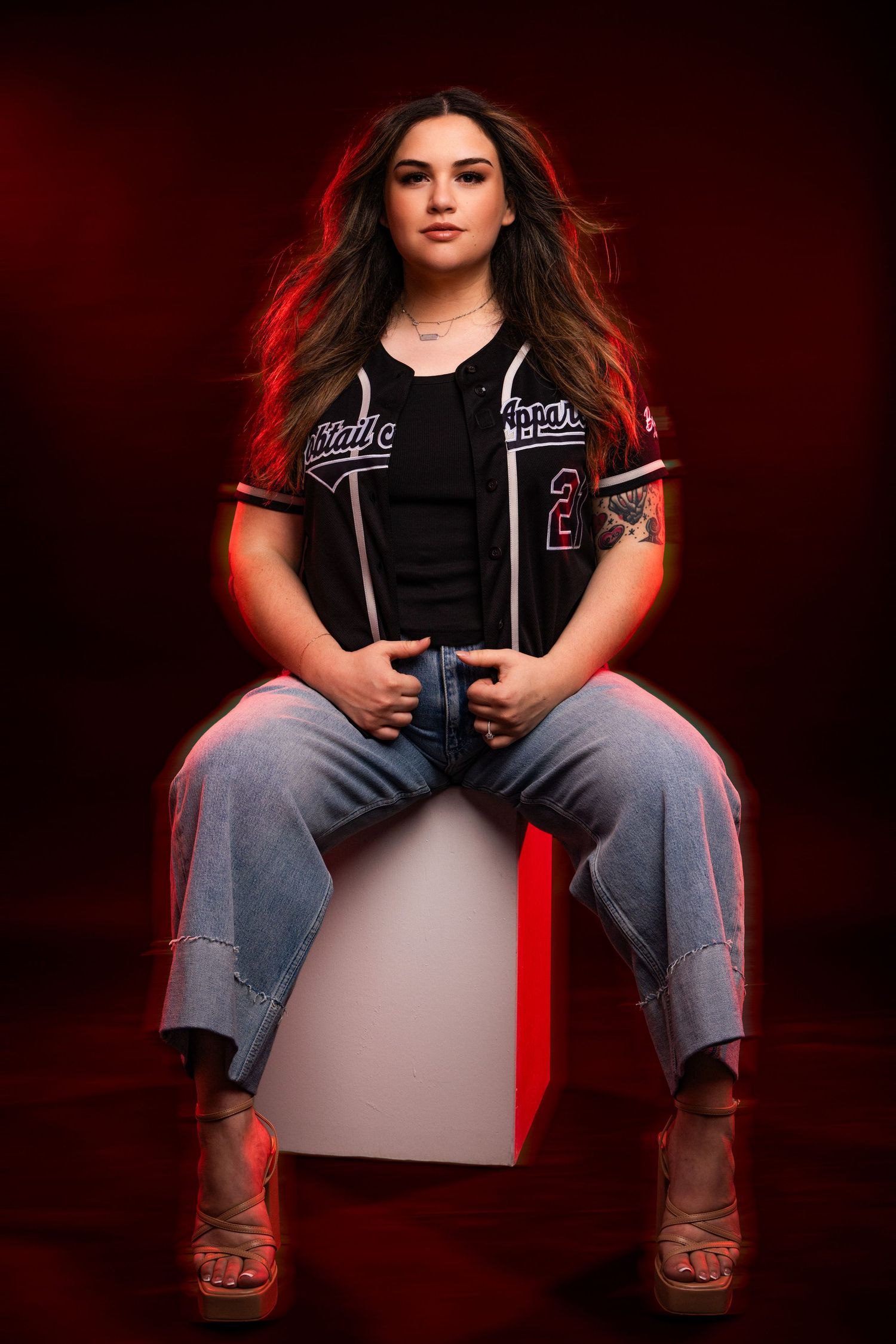 Dramatic portrait series with dark background showing someone seated on a red cube wearing jeans and black top.