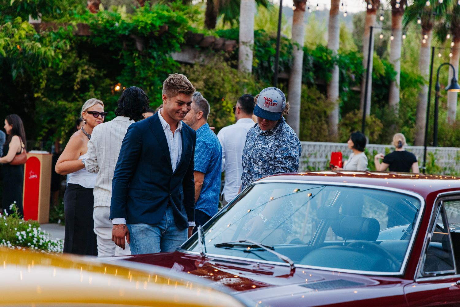 Classic vintage cars lined up at an outdoor event with palm trees and people gathering around.
