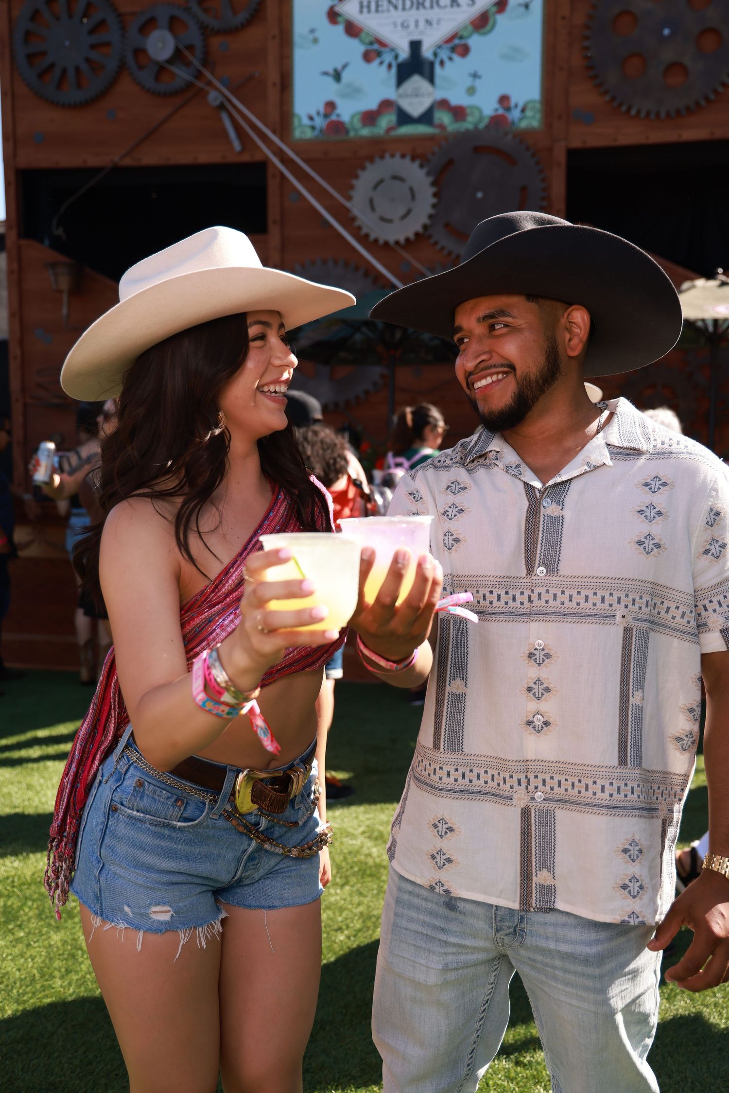 Couple in cowboy hats sharing drinks and laughing at an outdoor western-themed event with decorative gears in background.