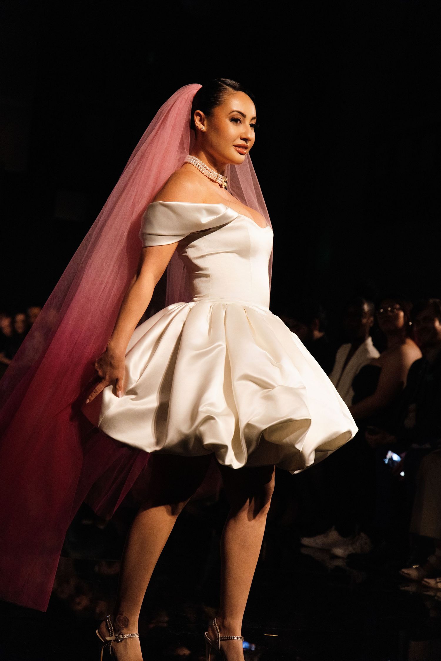 A model in a short white wedding dress with a pink veil walks the runway during a fashion show.