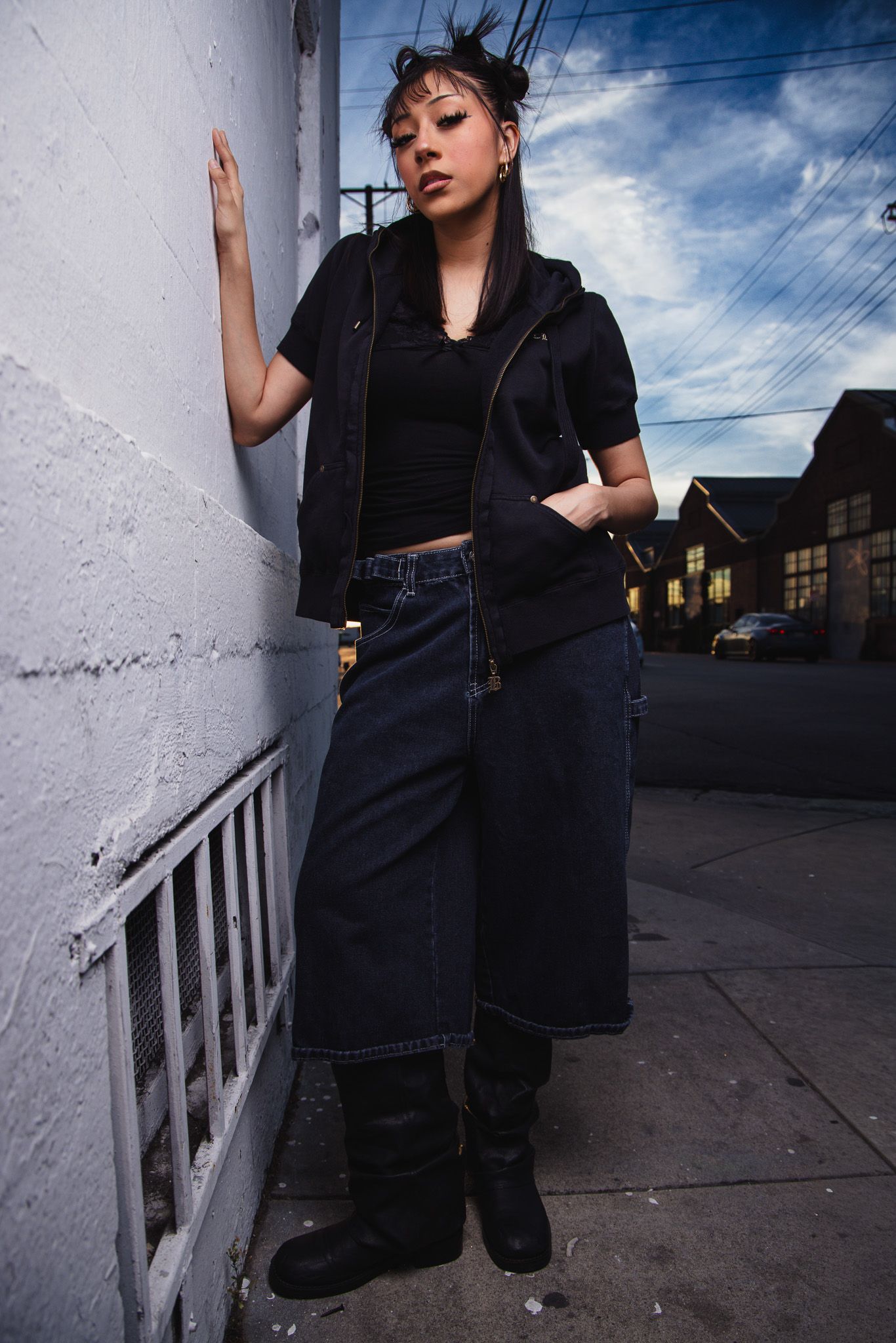 Fashion model in dark outfit poses against white wall and dramatic sky in urban setting.