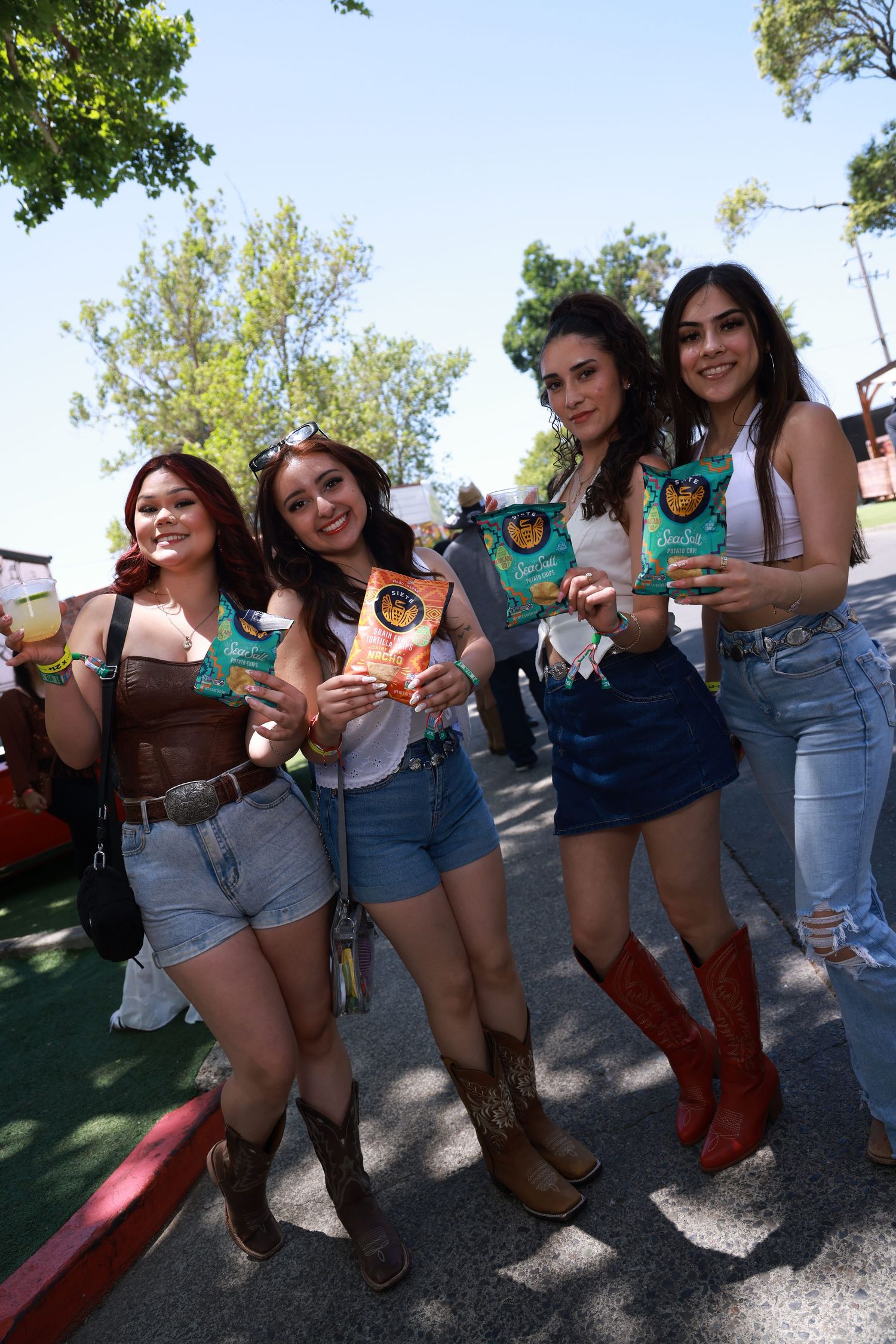 Four young women in cowboy boots and denim shorts pose together outdoors at a festival, holding snack bags.