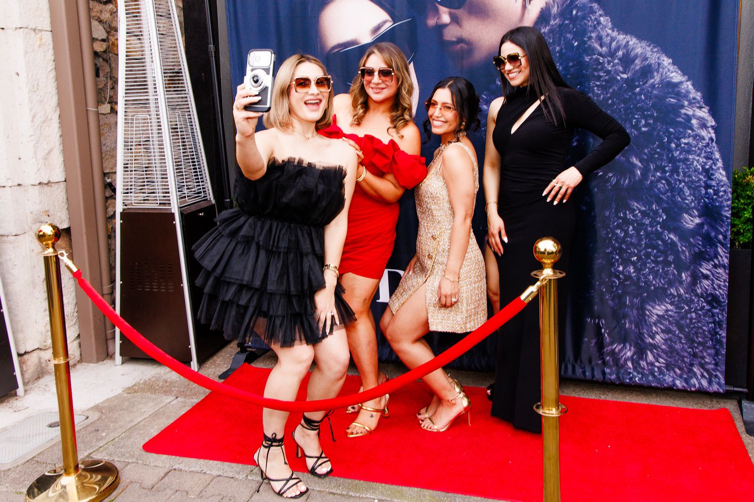 Four women posing for a selfie on a red carpet event, dressed in elegant outfits at a glamorous venue.