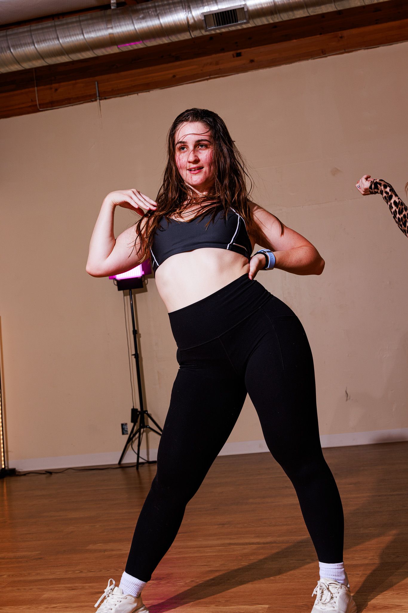 Woman in black sports bra and leggings dancing in a studio with wood floors.