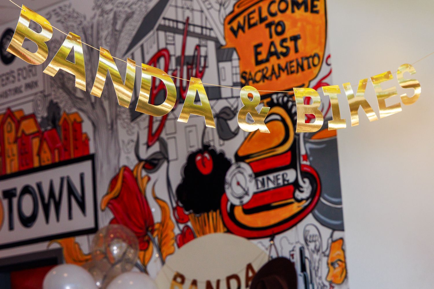 Banda & Bikes event banner with gold letters against East Sacramento mural backdrop with balloons.