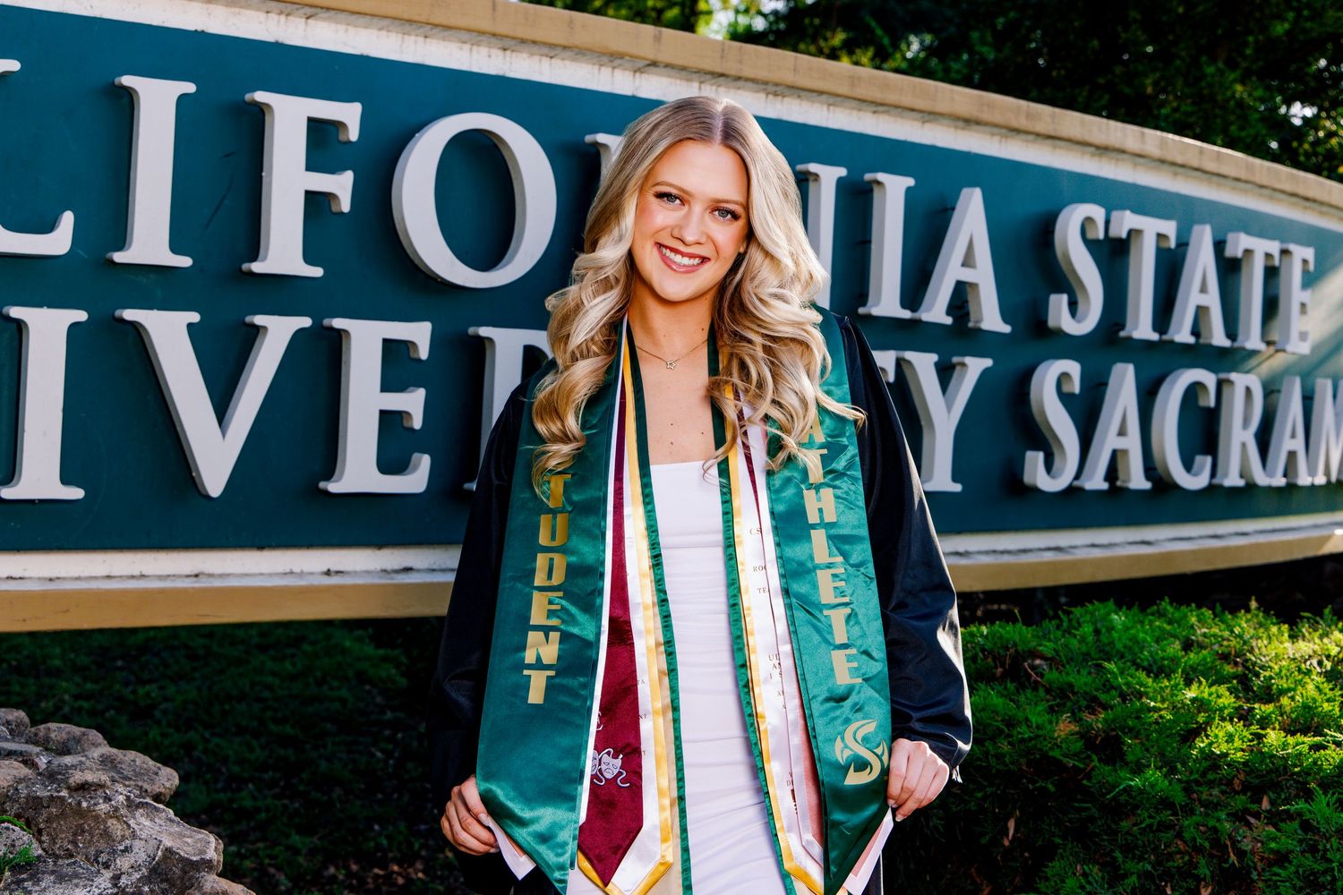 Graduate poses in cap and gown stoles at California State University Sacramento sign on a sunny day.