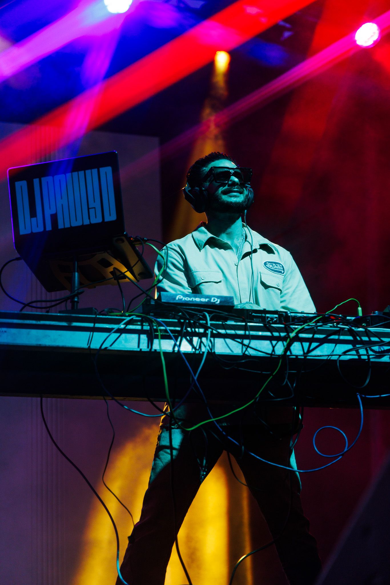 DJ Pradyo performing live on Pioneer DJ equipment under colorful stage lights with cables and a laptop display.