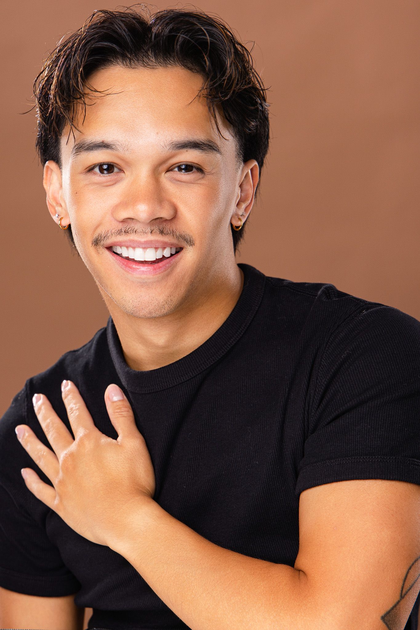 Smiling young man with mustache wearing black t-shirt poses with hand on chest against brown background.
