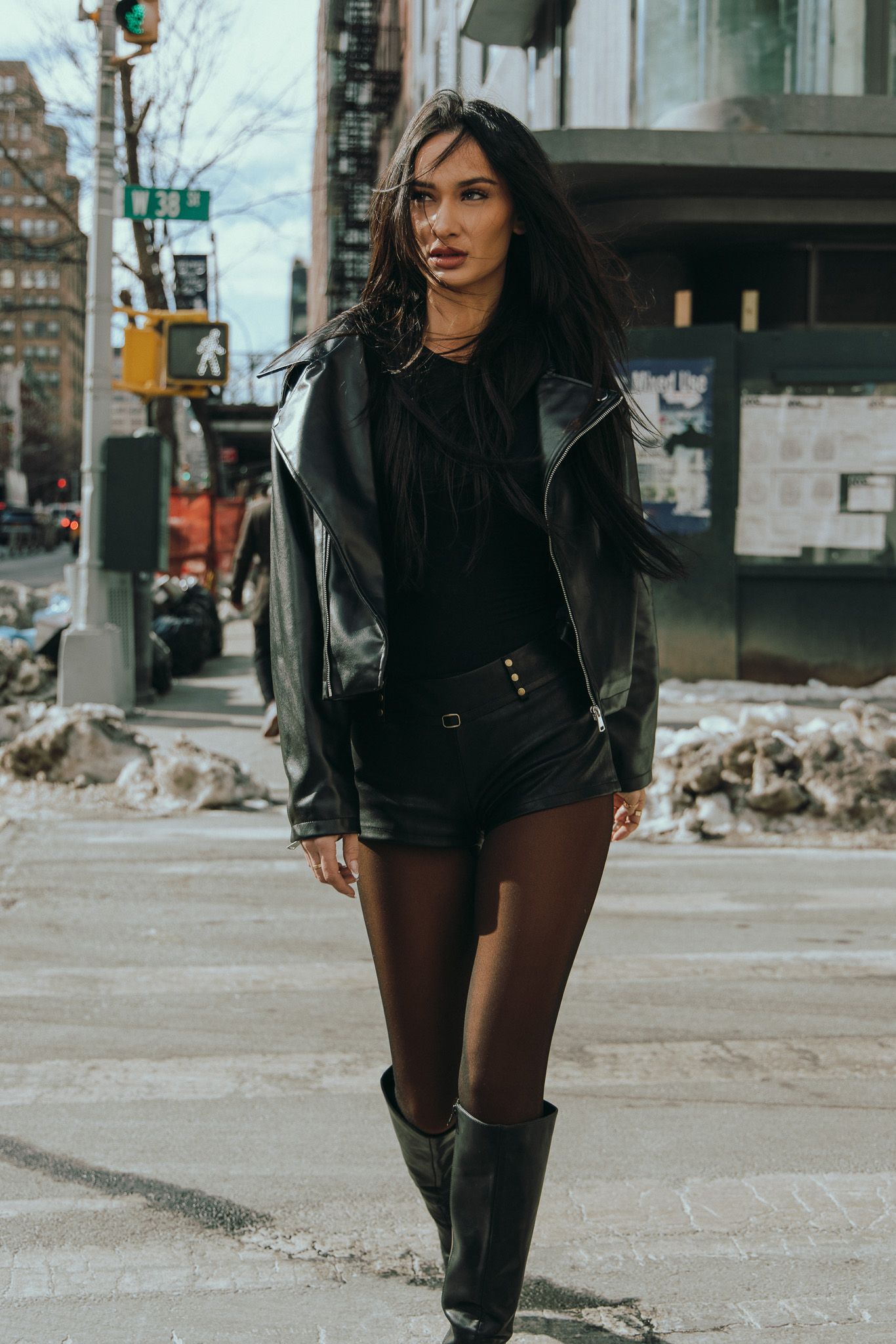 A stylish winter street fashion look featuring black thigh-high boots, shorts, and a dark coat on a snowy city sidewalk.