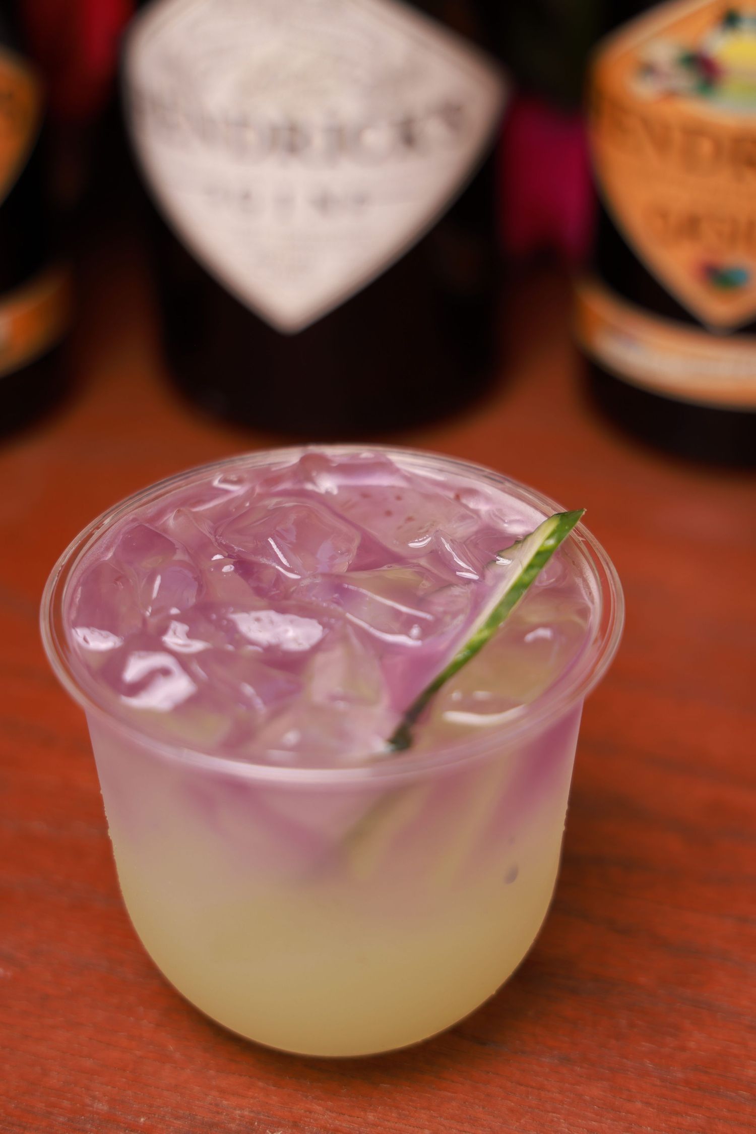 A purple-tinted cocktail with ice and garnish served in a rocks glass on a wooden surface.