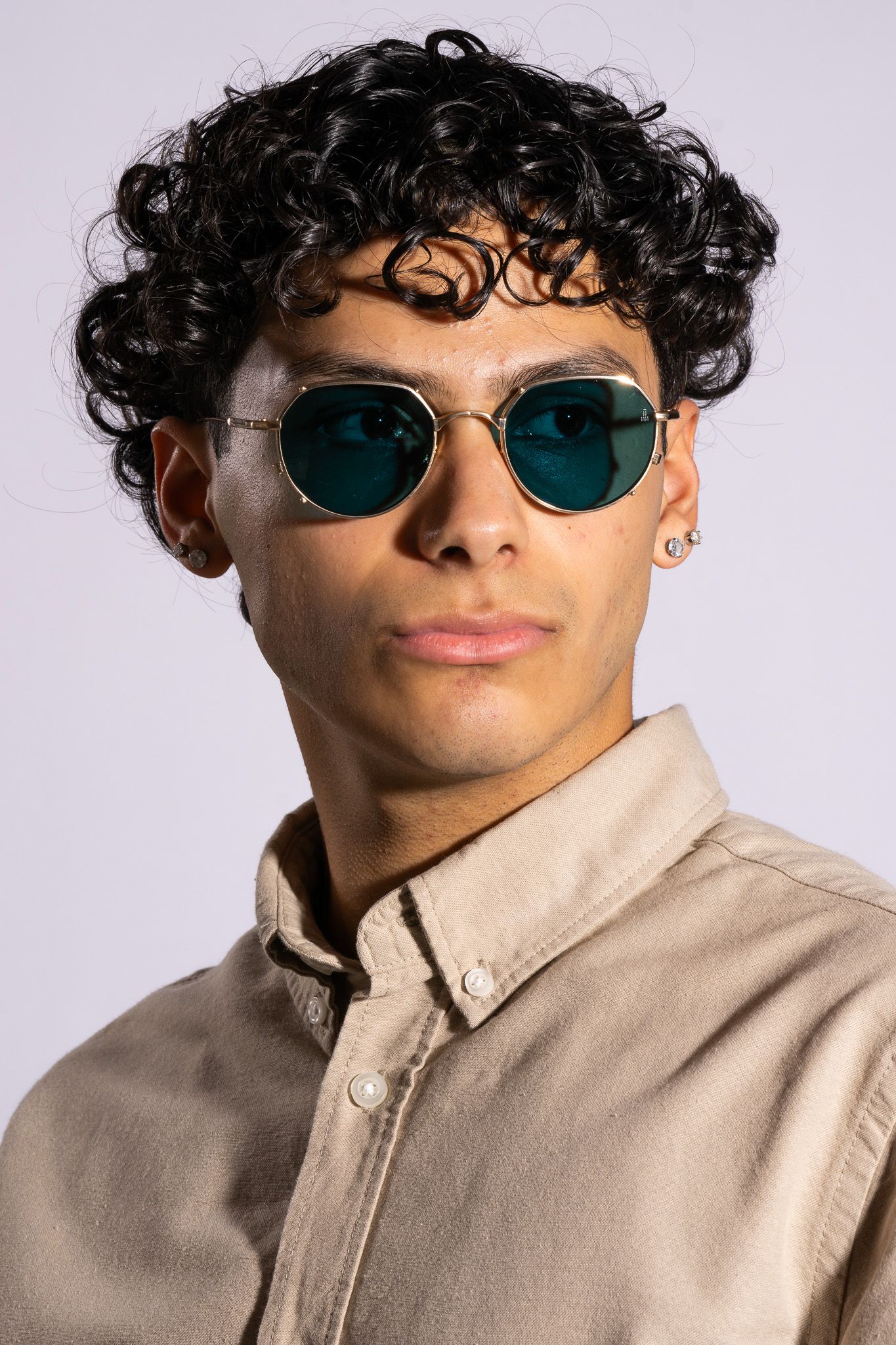 Young man with curly black hair wearing teal lens geometric sunglasses and beige button-up shirt.