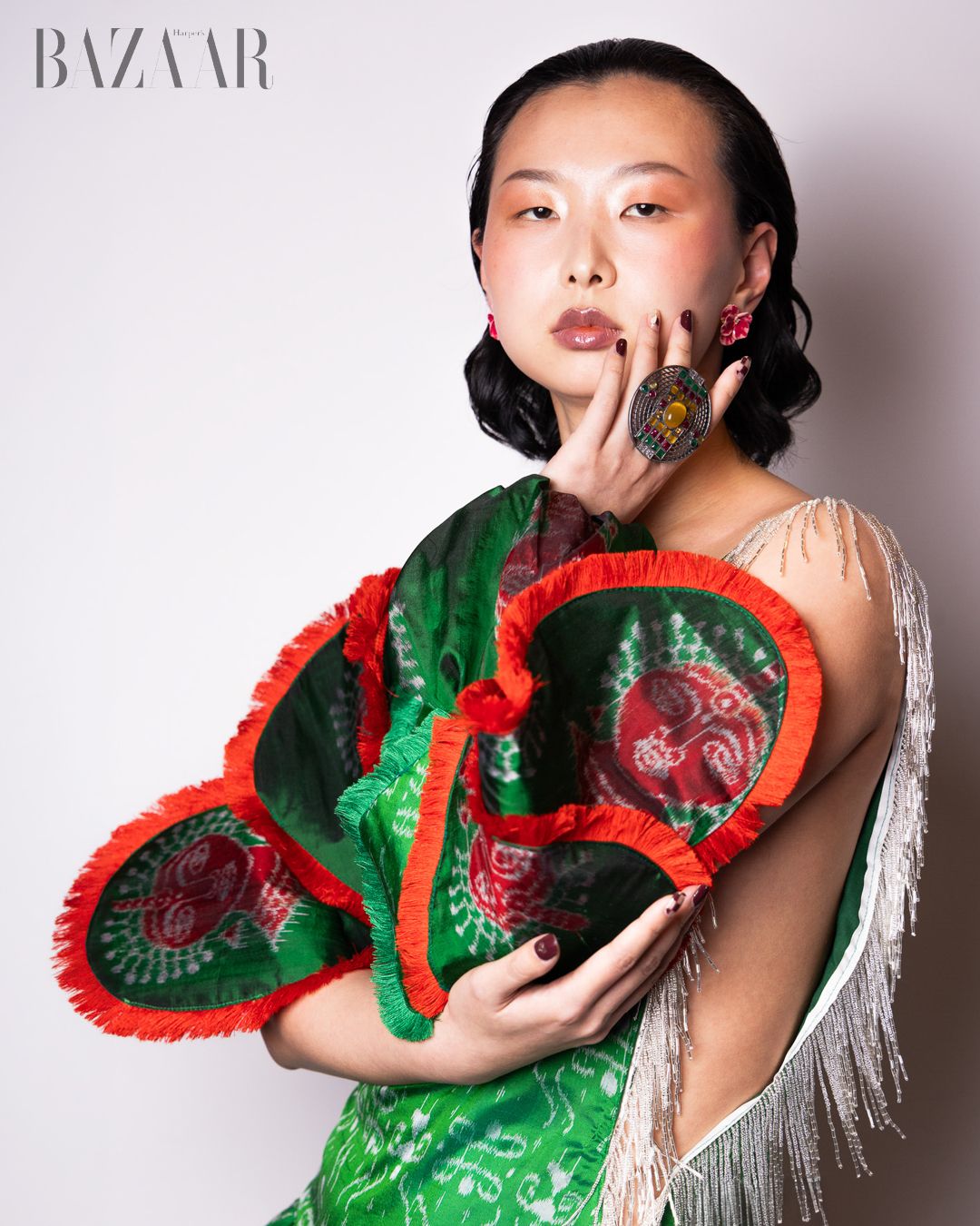 Model poses with green and red ruffled fan accessory, wearing fringe dress and floral earrings in Harper's Bazaar editorial.